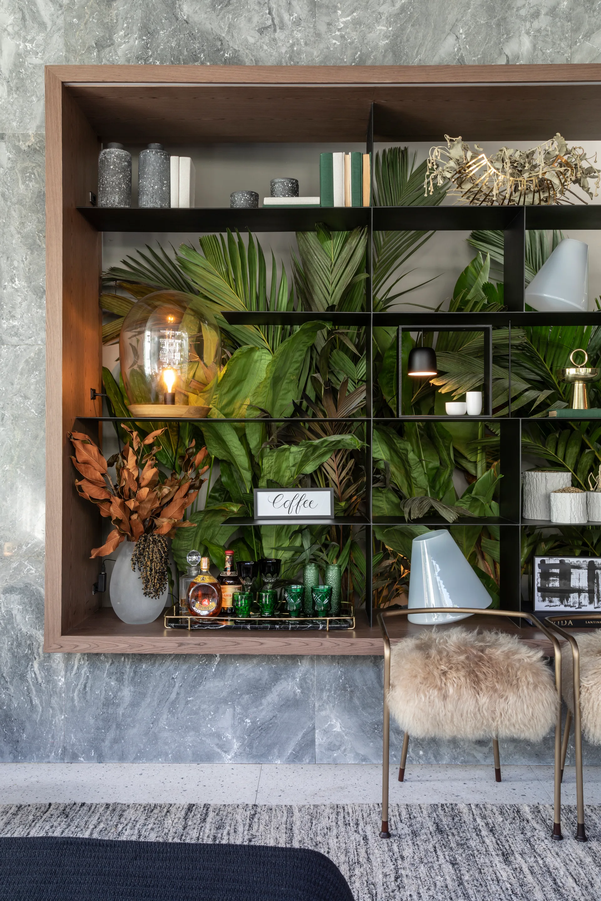 A wooden shelving unit recessed in a grey marble wall features thin shelves filled with decor and glassware against a backdrop of green palm leaves.