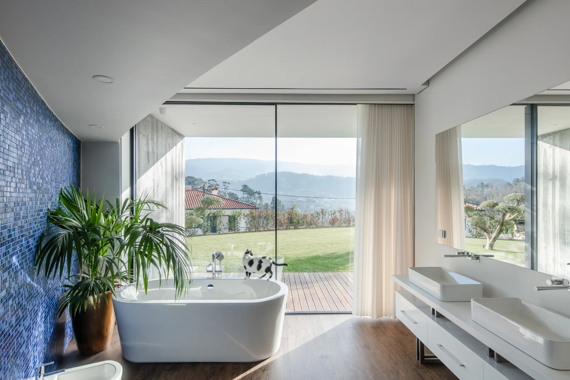 A modern bathroom features a white freestanding bathtub next to a glass wall, a blue mosaic tile wall, and a white double vanity.