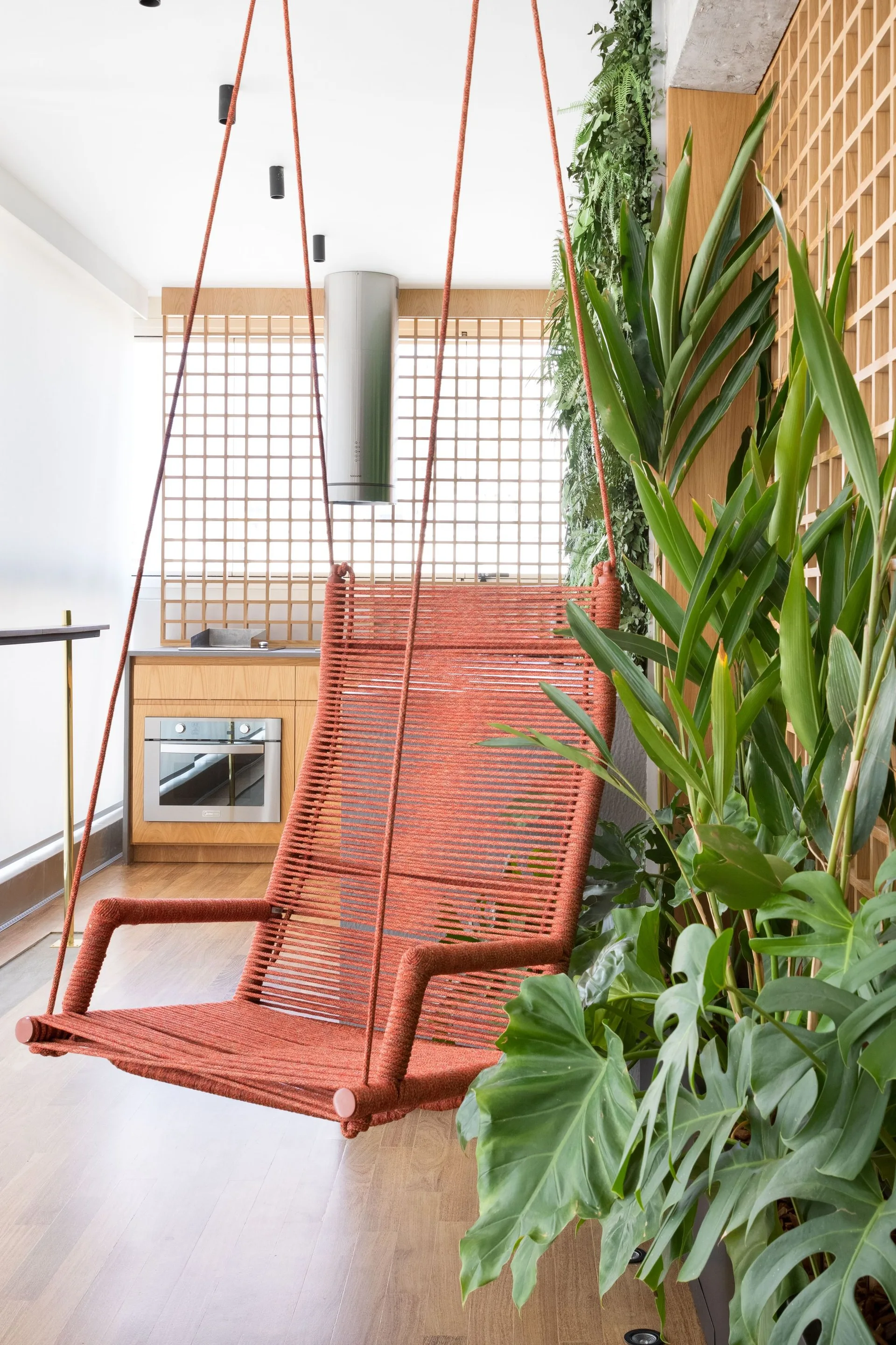 A modern balcony features an orange rope hanging chair suspended next to leafy green plants, wood paneling, and an outdoor kitchenette.