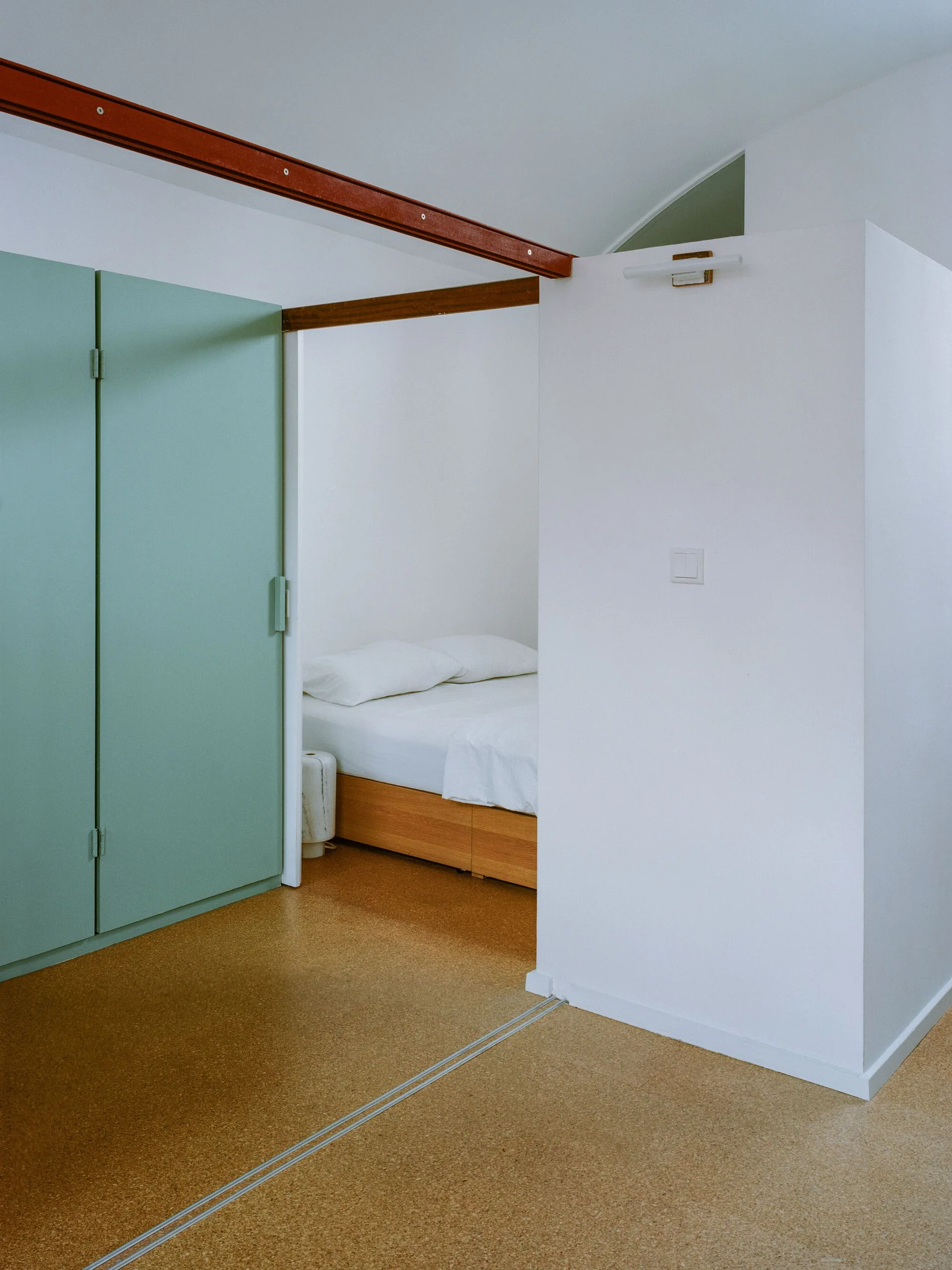 A minimalist bedroom space features a wooden bed with white linens, a prominent mint green closet door, and speckled cork flooring.