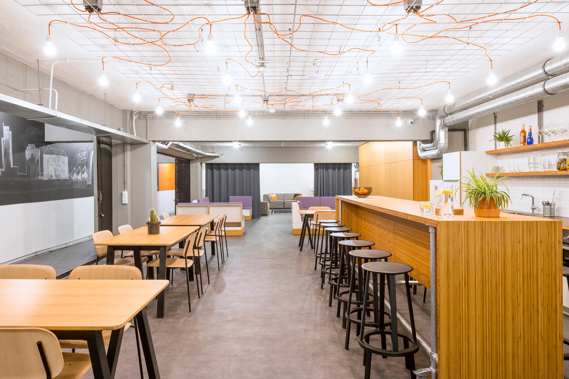 A spacious industrial dining room featuring long wooden tables, black bar stools, concrete floors, and a white ceiling grid with exposed orange wiring.
