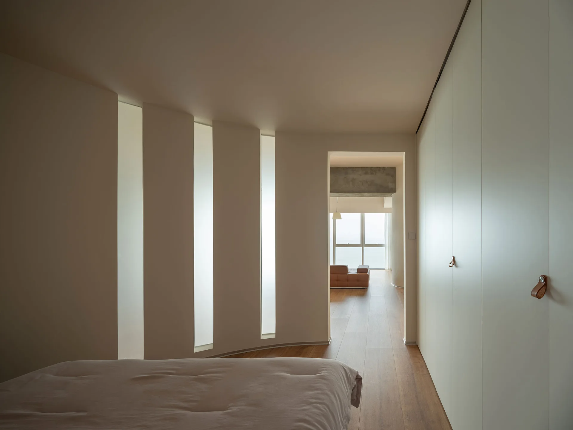 A minimalist bedroom features vertical wall light panels, a floor-to-ceiling closet with leather pulls, and a doorway looking into a living room.