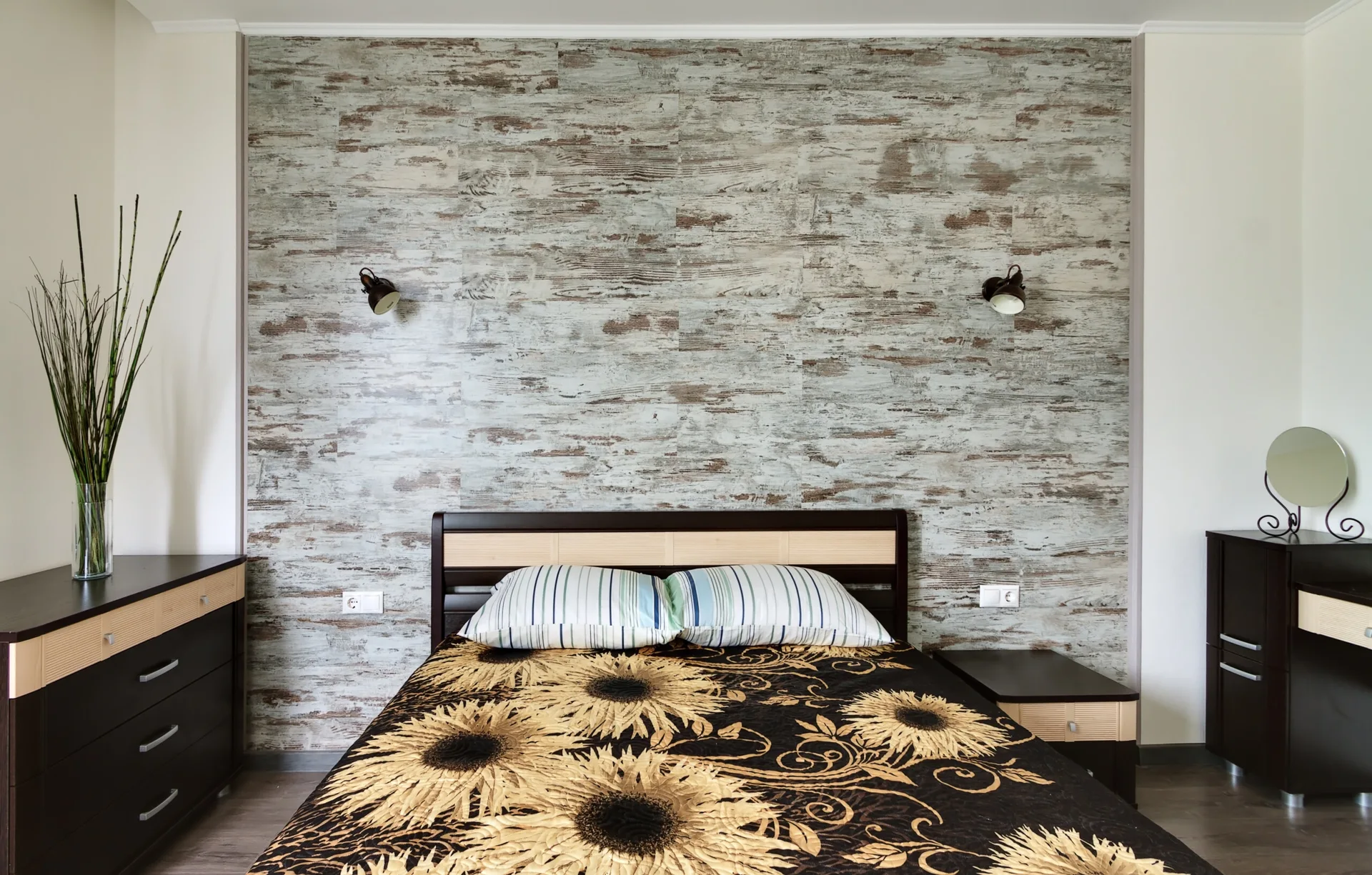 A bedroom featuring a bed with sunflower bedding set against a weathered wood-look accent wall, flanked by dark wood dressers and wall sconces.