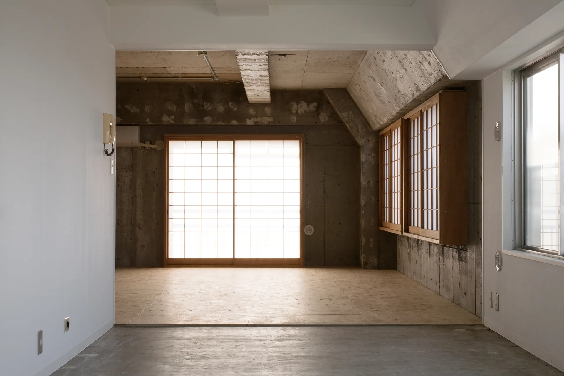 A minimalist room featuring raw concrete walls, a raised plywood floor platform, and glowing sliding shoji screens.