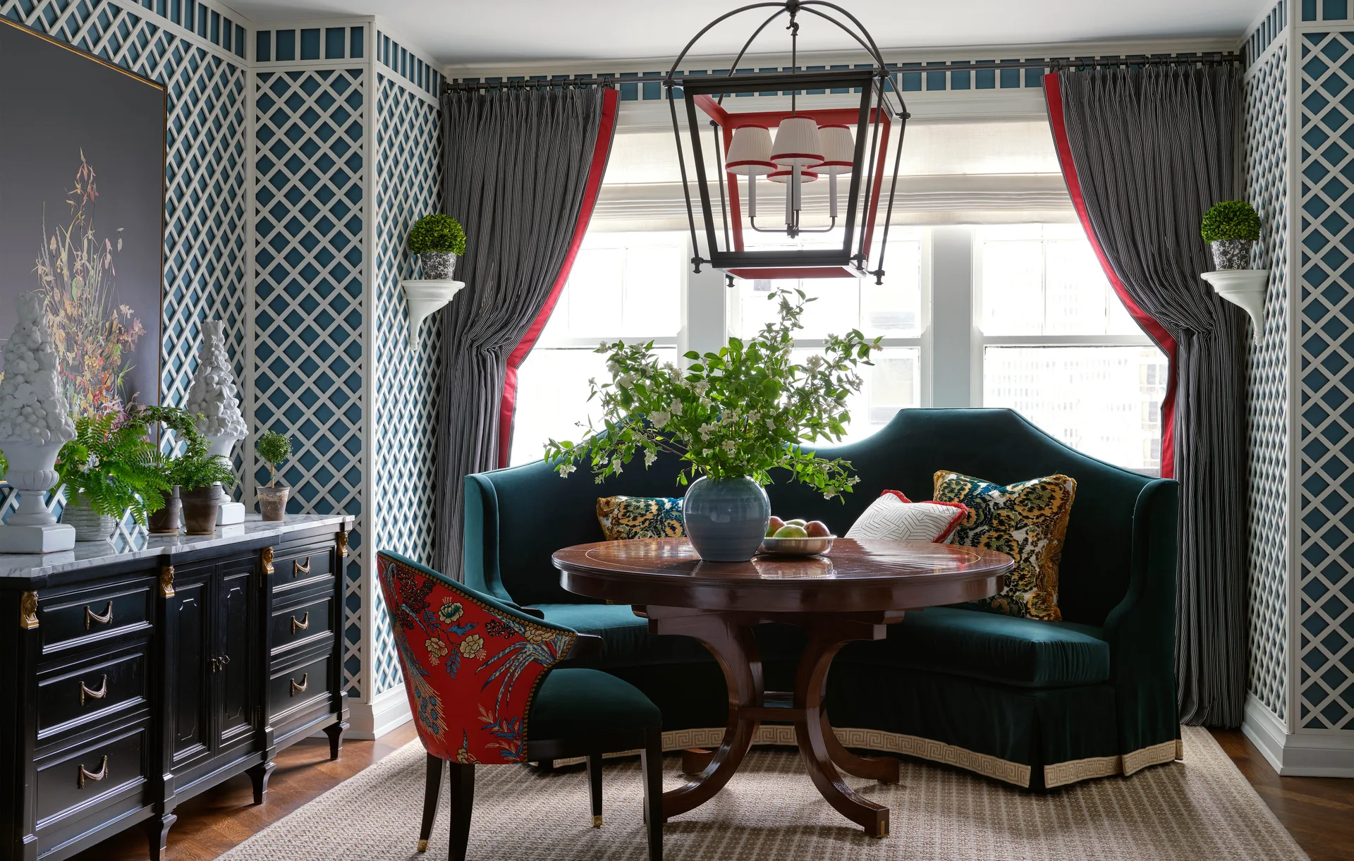 A dining nook features a curved dark green velvet banquette around a round wood table against blue trellis wallpaper and a large window.