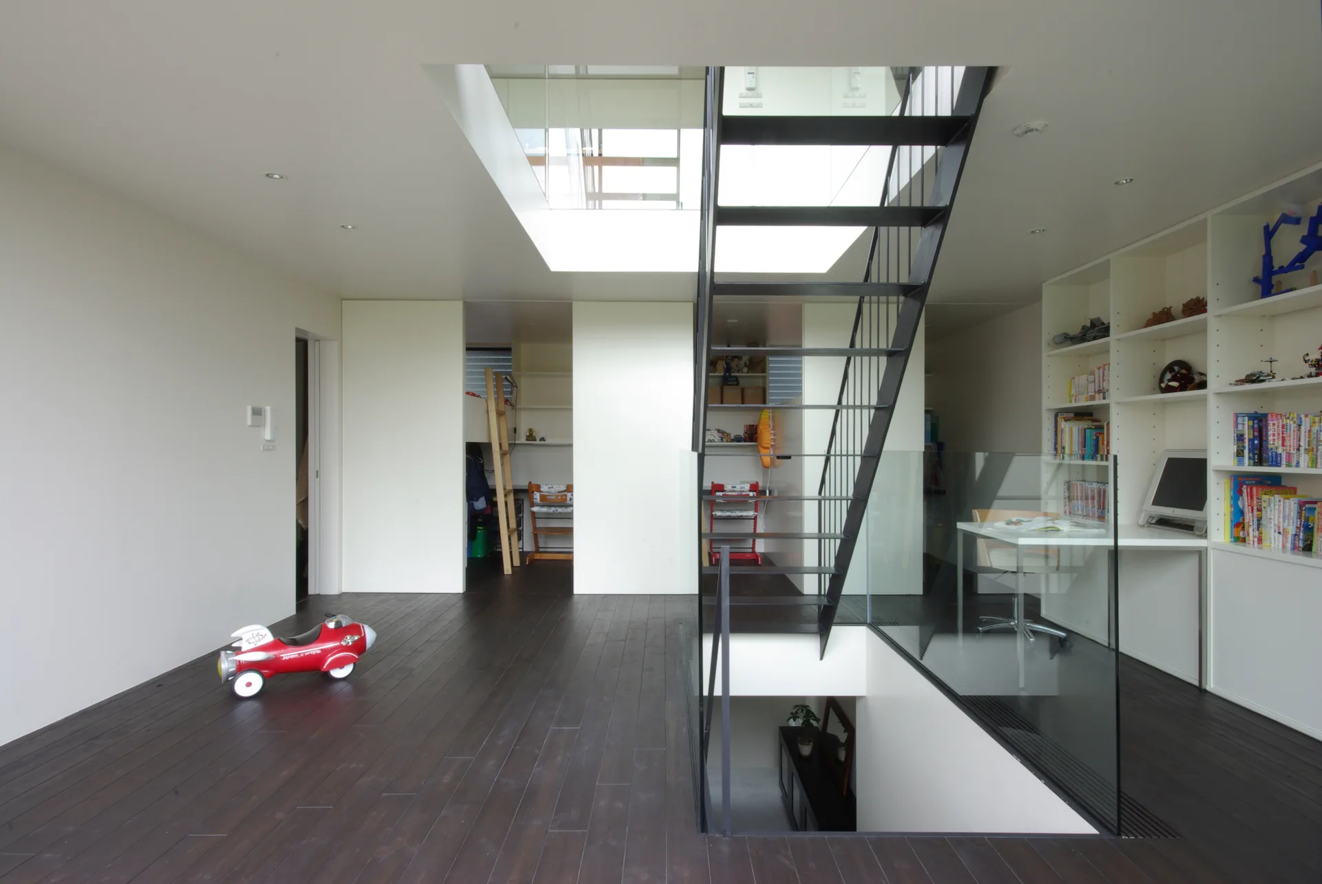 A minimalist home interior features a central black metal open-riser staircase with a glass railing, dark wood floors, and white built-in shelving.