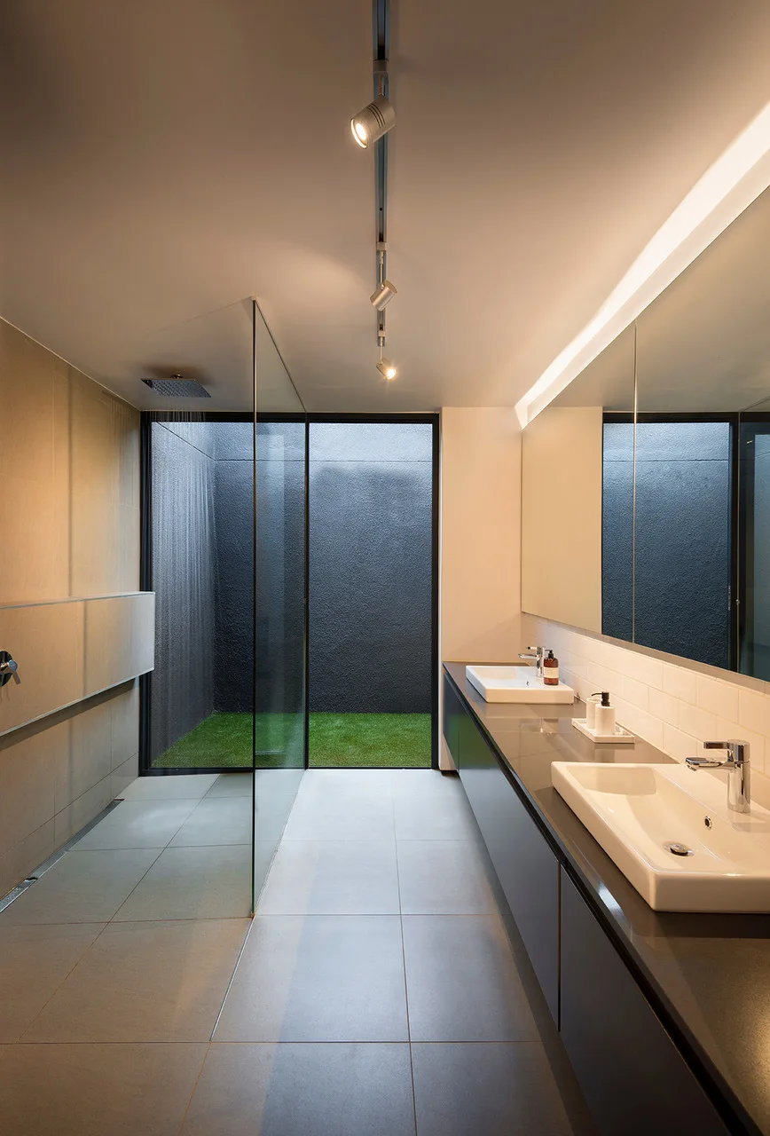 A minimalist bathroom features a long dark gray vanity with two sinks, a glass-walled shower, and a large window looking onto a small green courtyard.