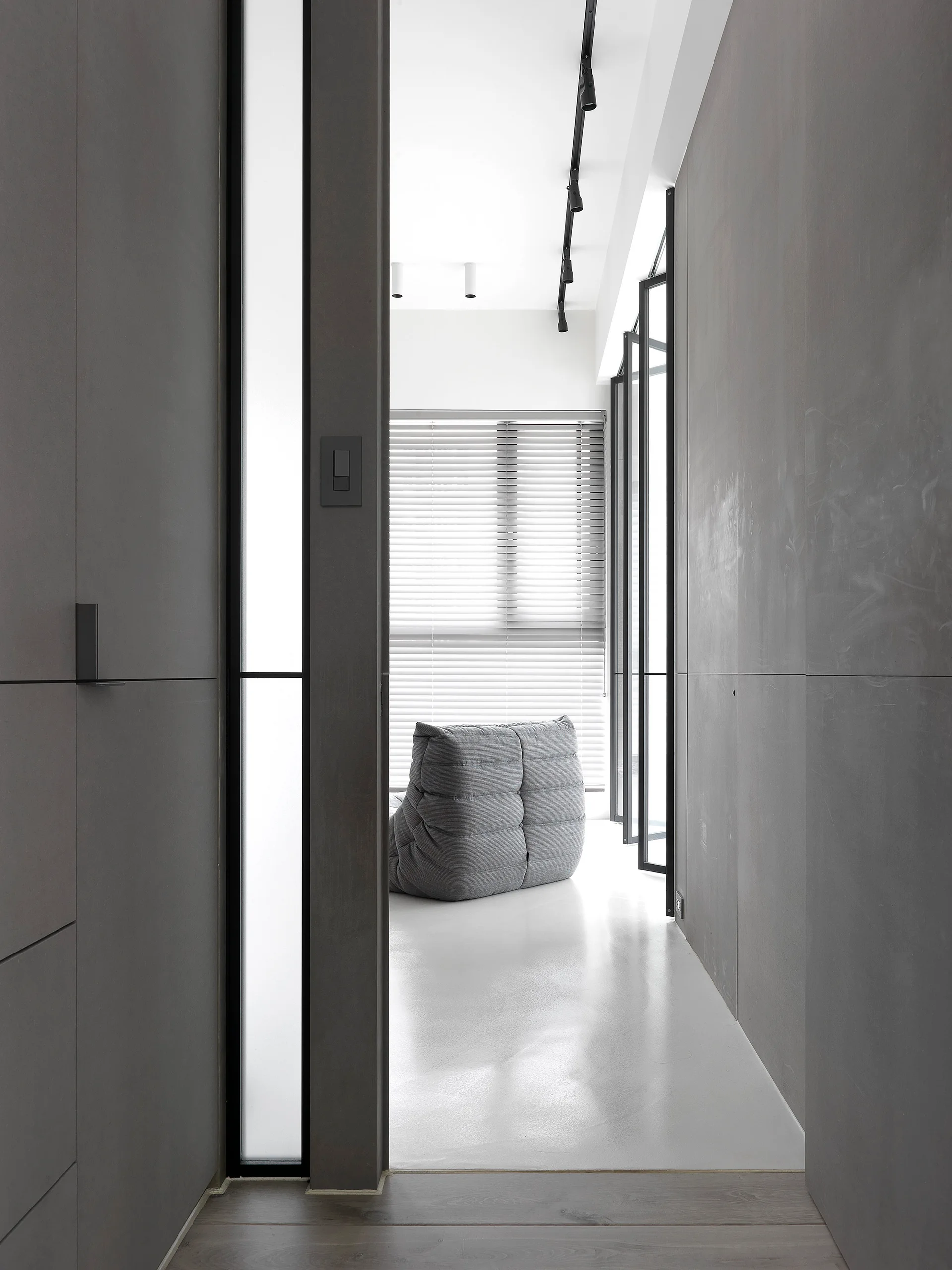 A minimalist entryway with matte grey paneled walls opens into a bright living room featuring a quilted grey lounge chair and venetian blinds.
