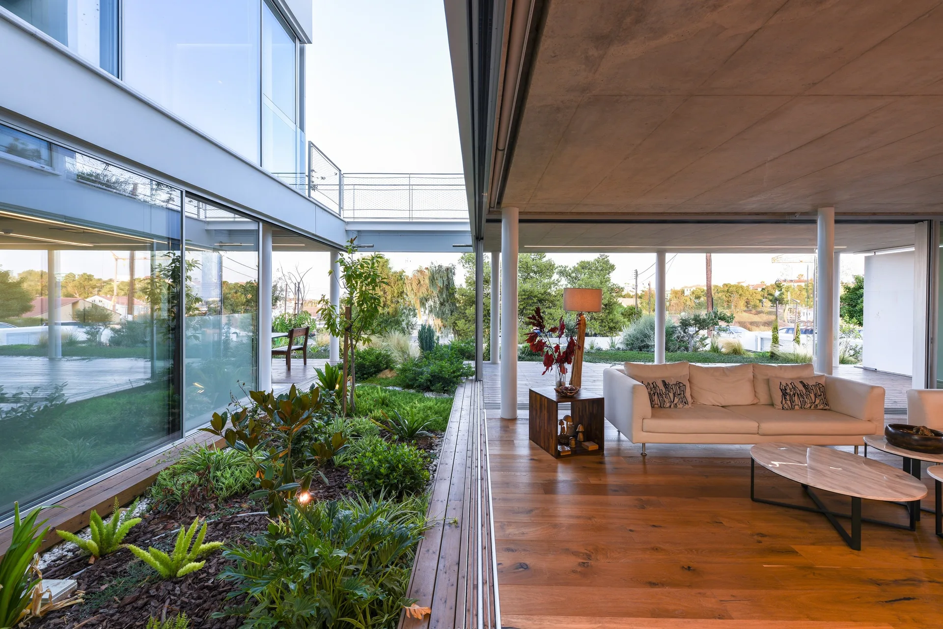 A modern living room features a white sofa, wood floors, and an exposed concrete ceiling facing an indoor garden courtyard through glass walls.