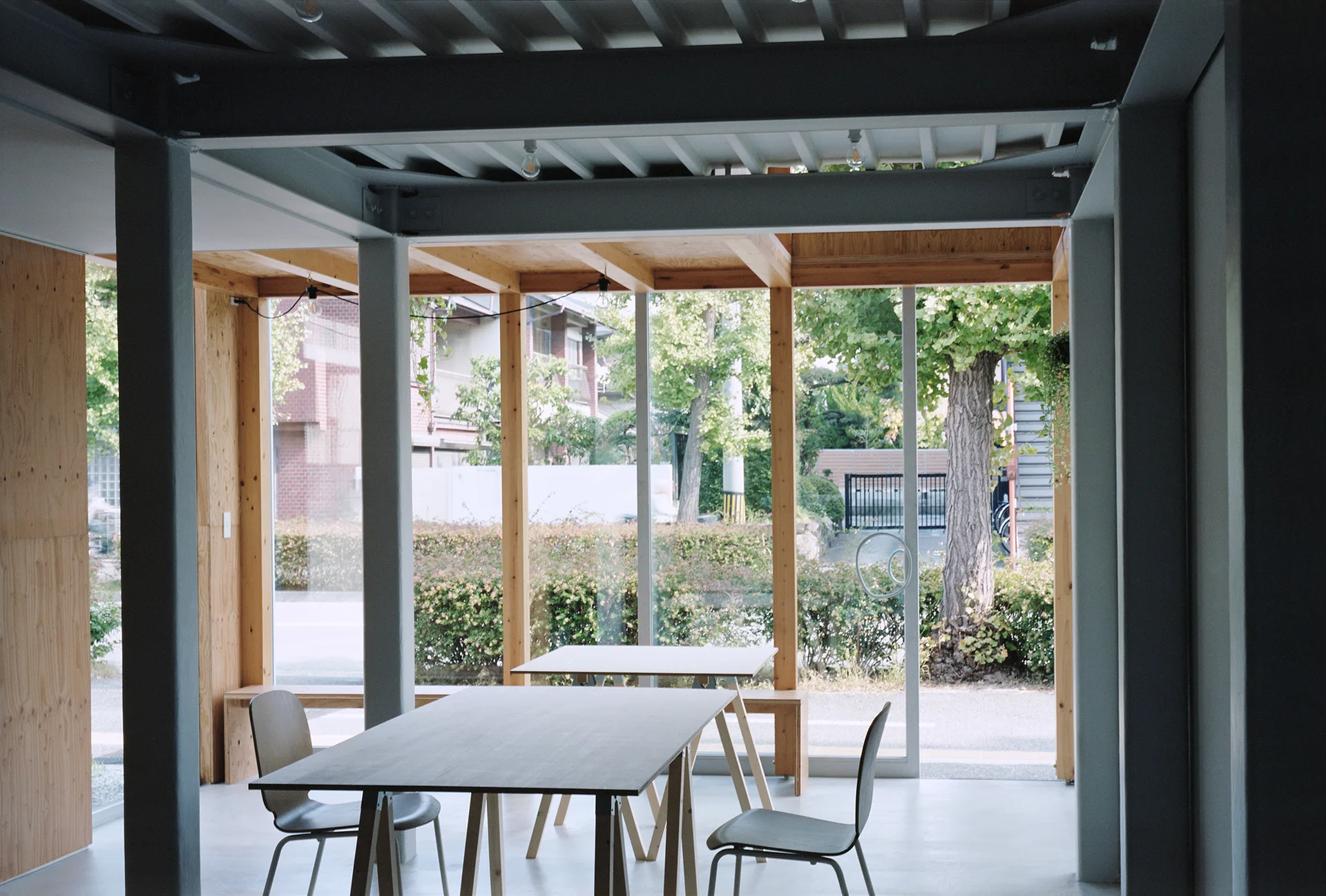 A minimalist dining area with exposed steel ceiling beams, light wood plywood walls, and simple wood tables and chairs next to large windows.