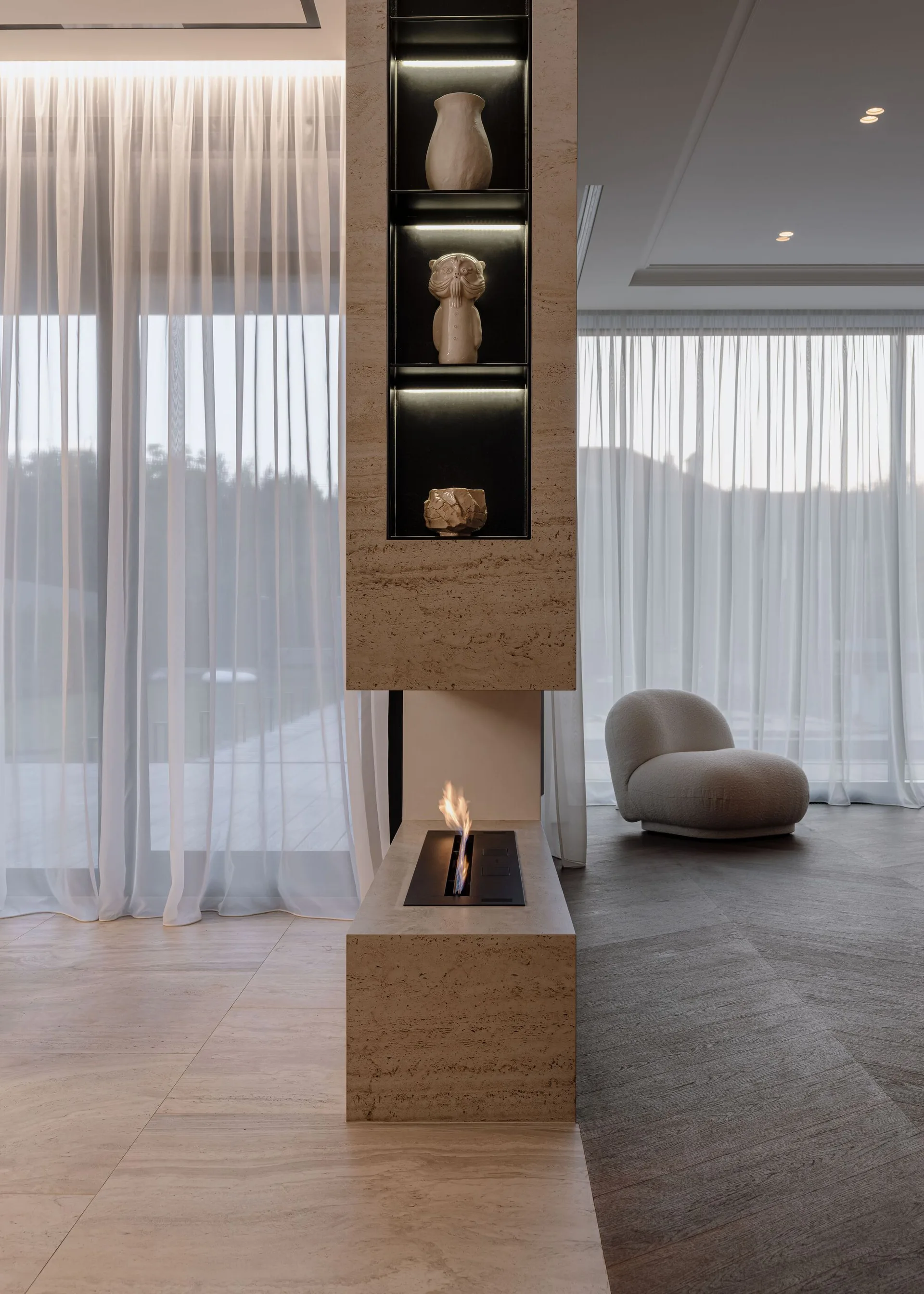 A modern stone column fireplace with integrated display niches divides a living space with sheer curtains and a curved lounge chair.