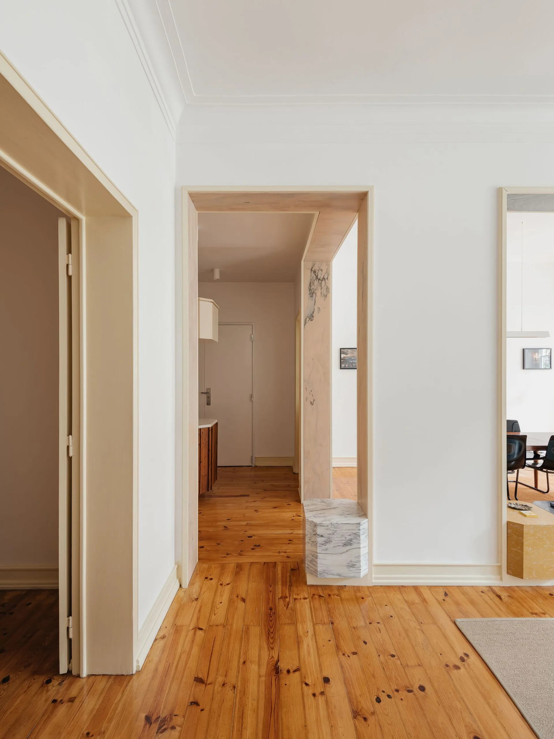 A minimalist white hallway features natural pine wood floors and custom wood-lined door frames with a hexagonal marble block at one base.