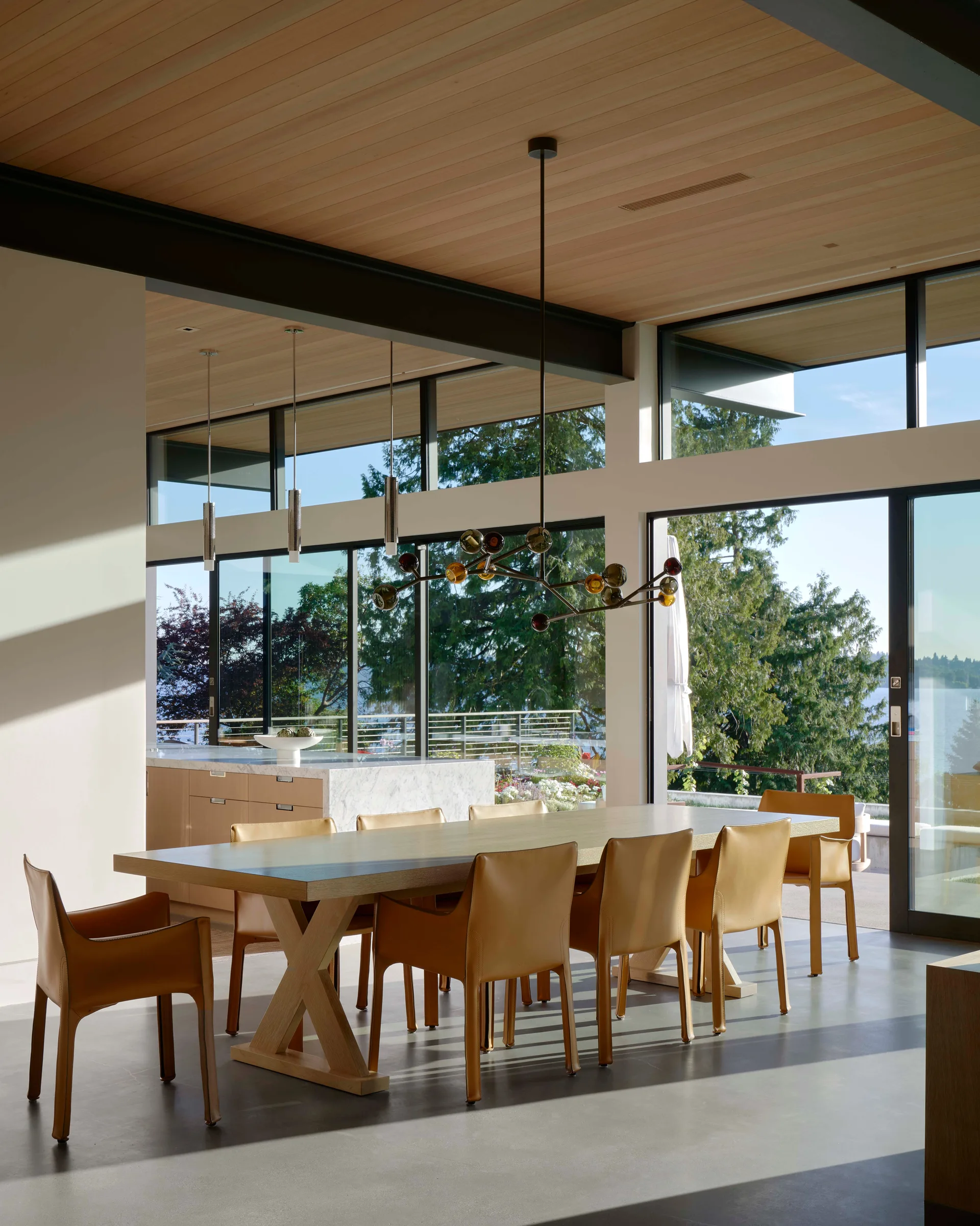 A long wooden dining table and tan leather armchairs sit under a wood-paneled ceiling next to large glass doors looking out at trees.