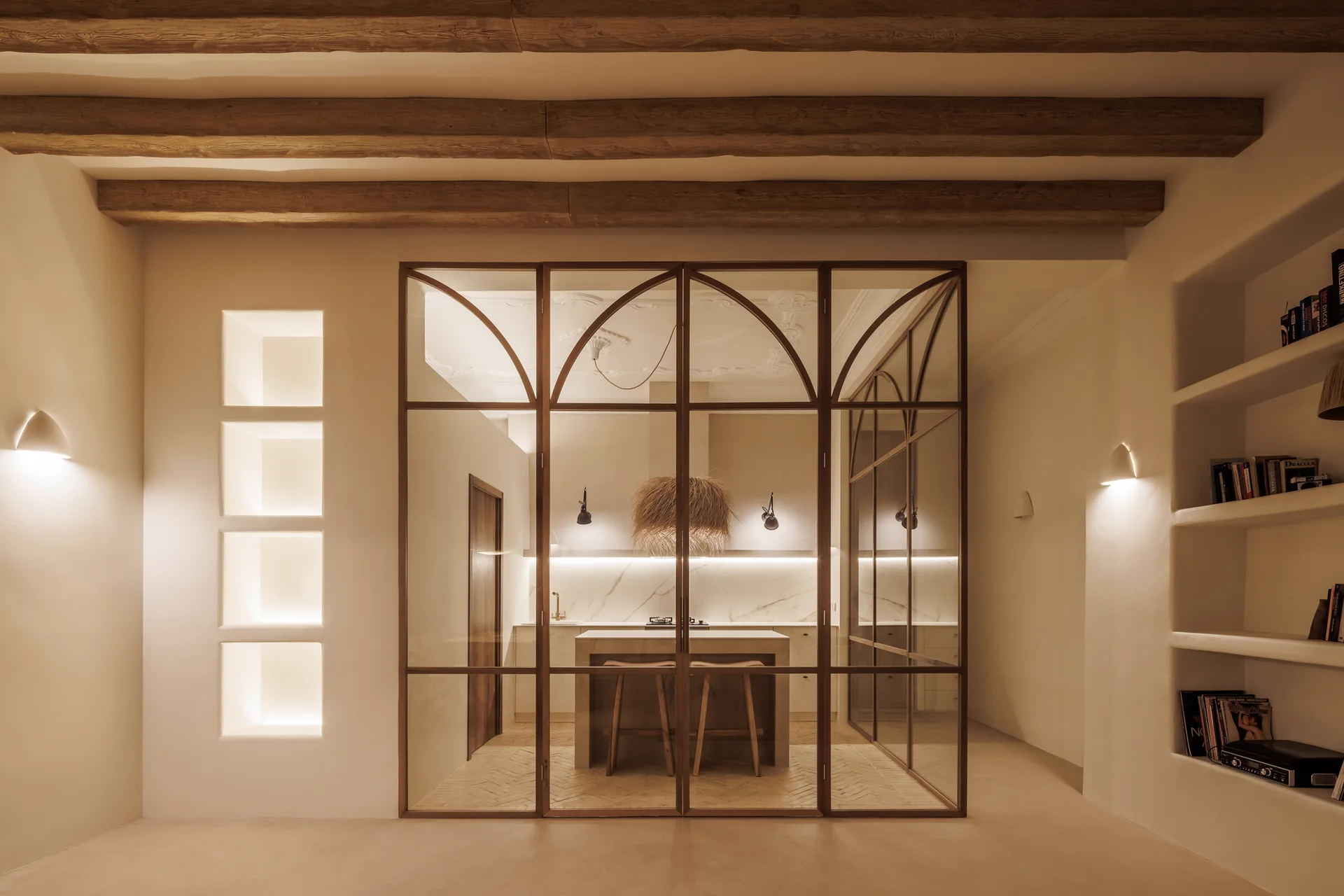 A modern kitchen enclosed by an arched metal and glass partition, featuring warm plaster walls, exposed wooden ceiling beams, and built-in niches.