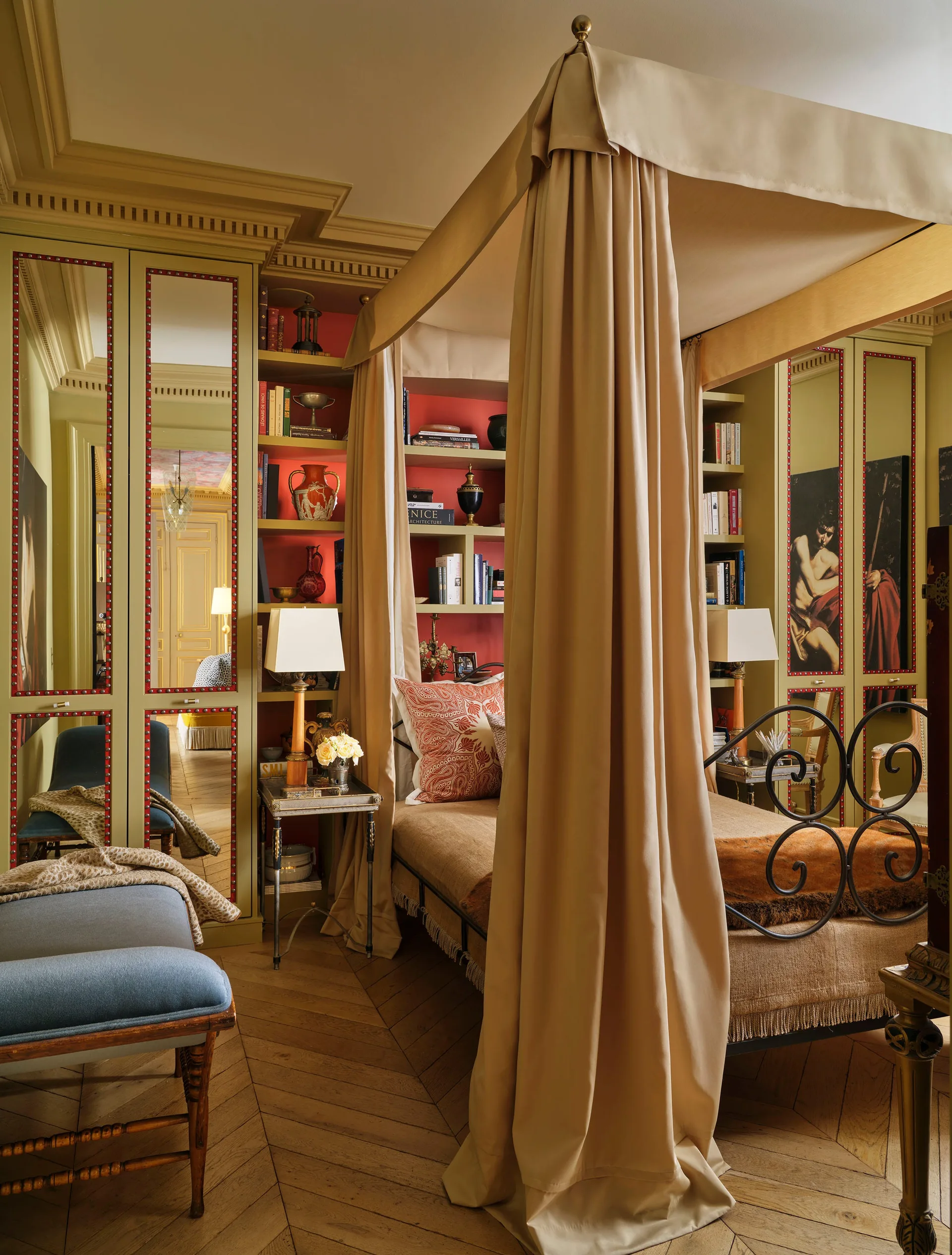 A traditional bedroom features a beige fabric canopy bed flanked by olive green built-in shelves with red backings and mirrored wardrobe doors.