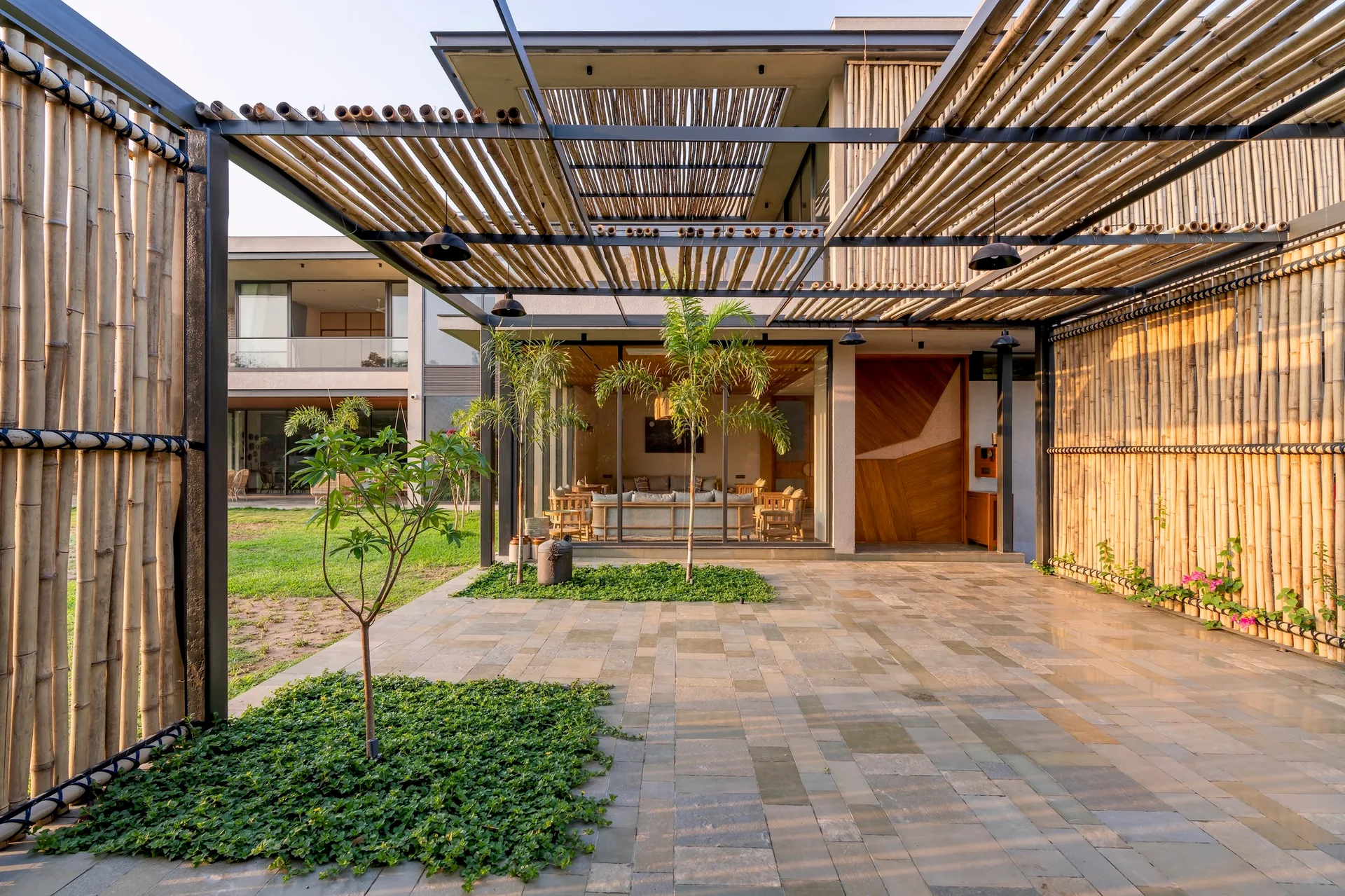 An outdoor patio features a large bamboo pergola, stone pavers with small garden cutouts, and glass walls looking into a living room.