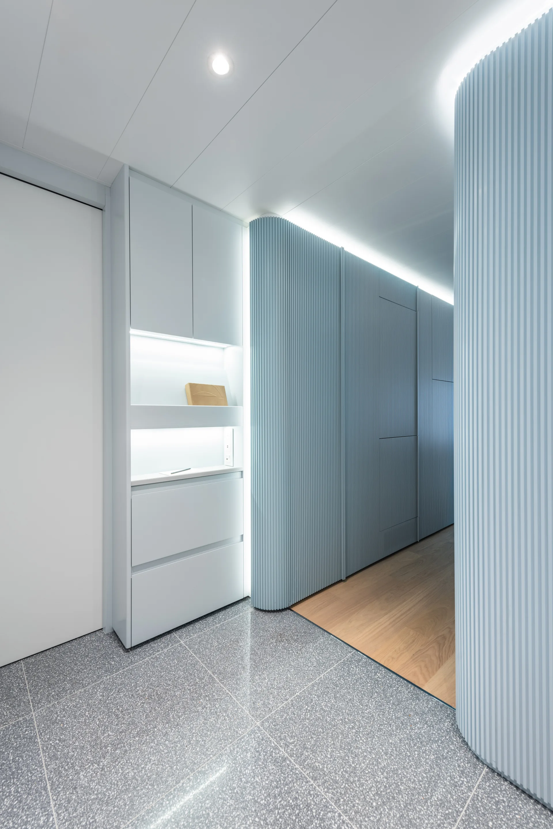 A modern hallway features light blue fluted wall panels, integrated cove lighting, and a transition from grey terrazzo tile to light wood flooring.