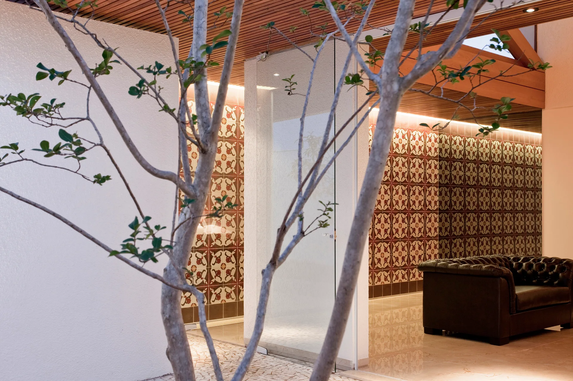 A tufted leather sofa sits near a geometric patterned tile wall and a glass partition under a slatted wood ceiling.