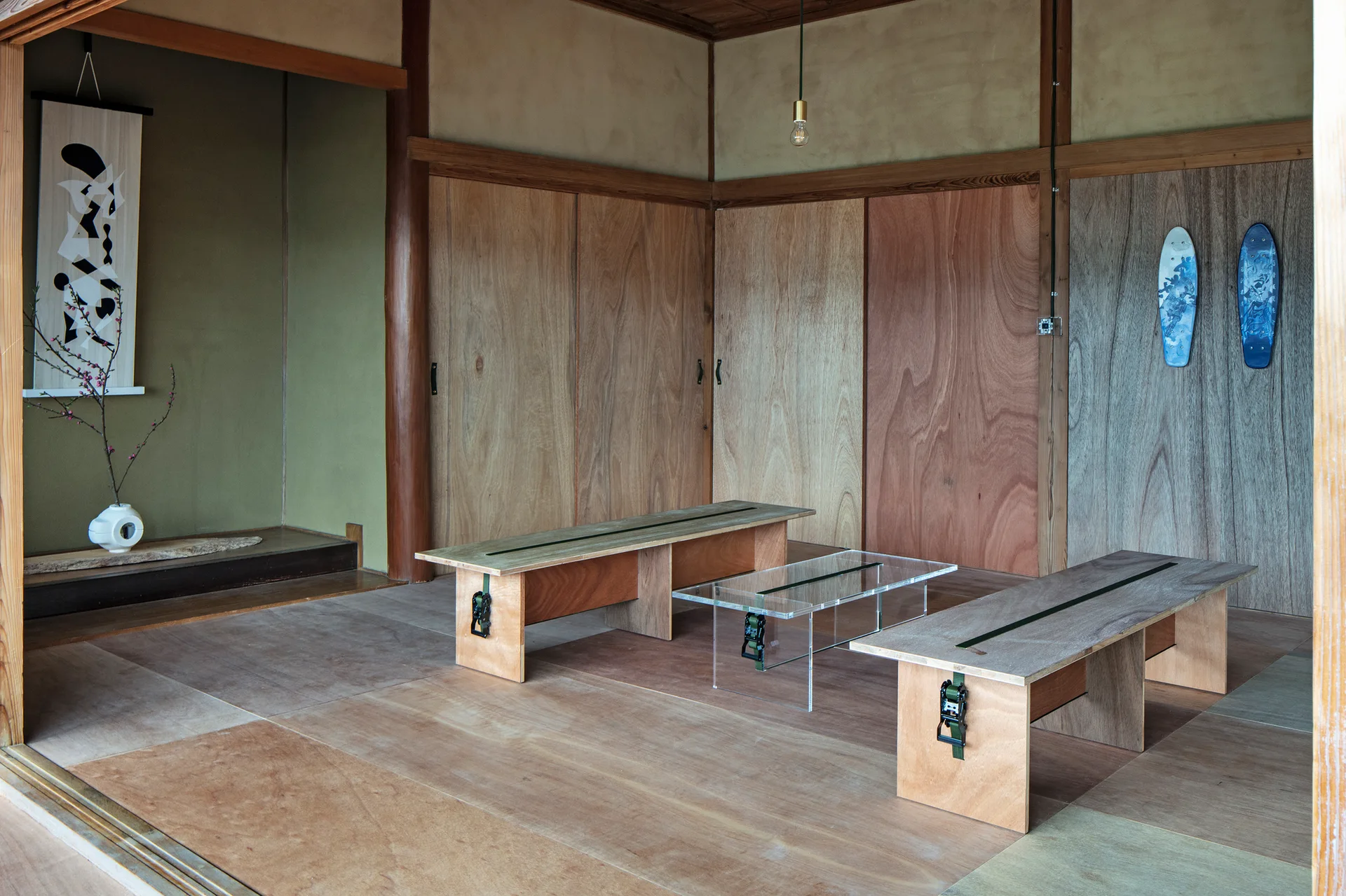 A minimalist wooden room features two plywood benches with green ratchet straps and a clear acrylic coffee table.