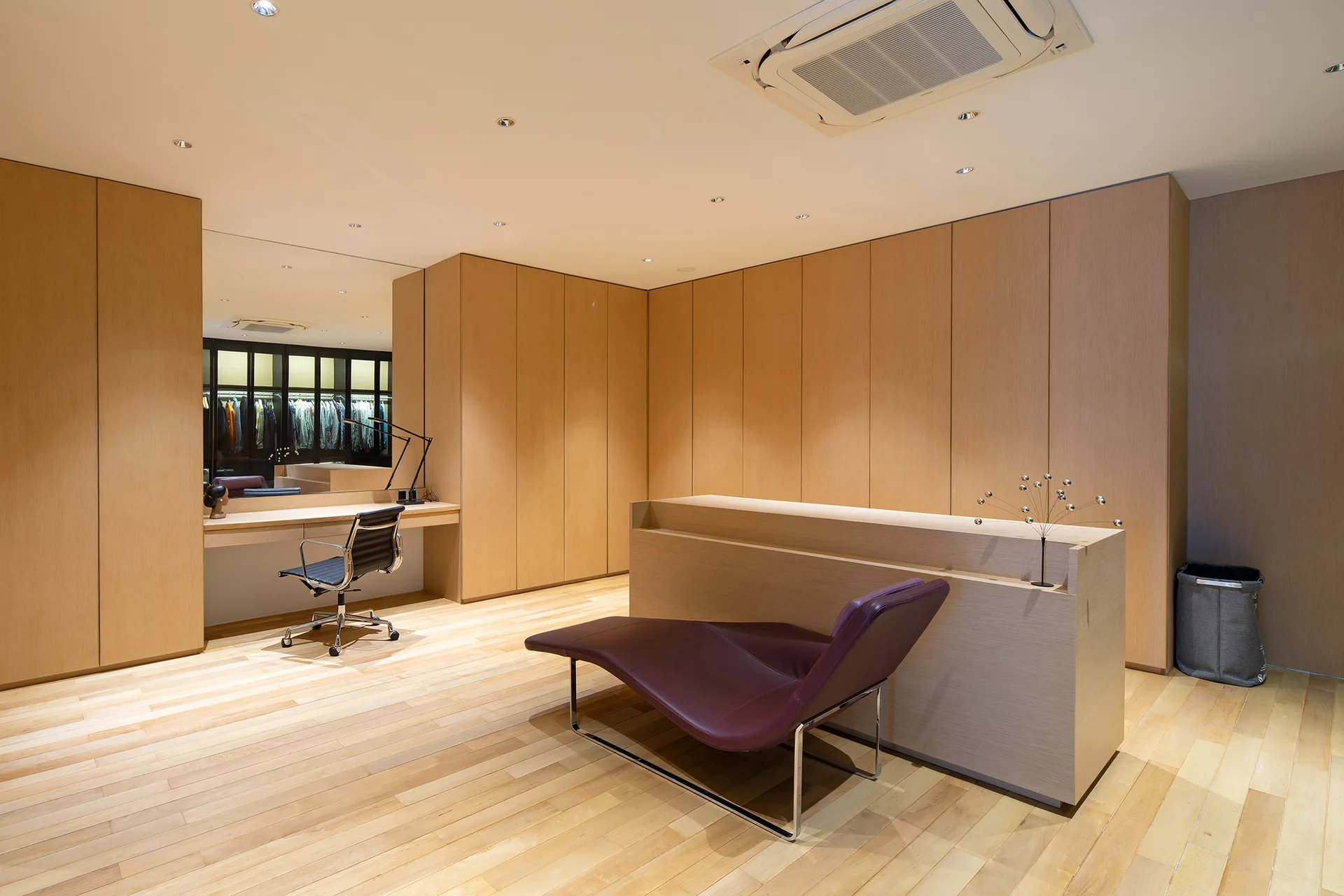 A minimalist dressing room with light wood paneled cabinetry, a built-in vanity station, a black desk chair, and a central purple leather chaise.