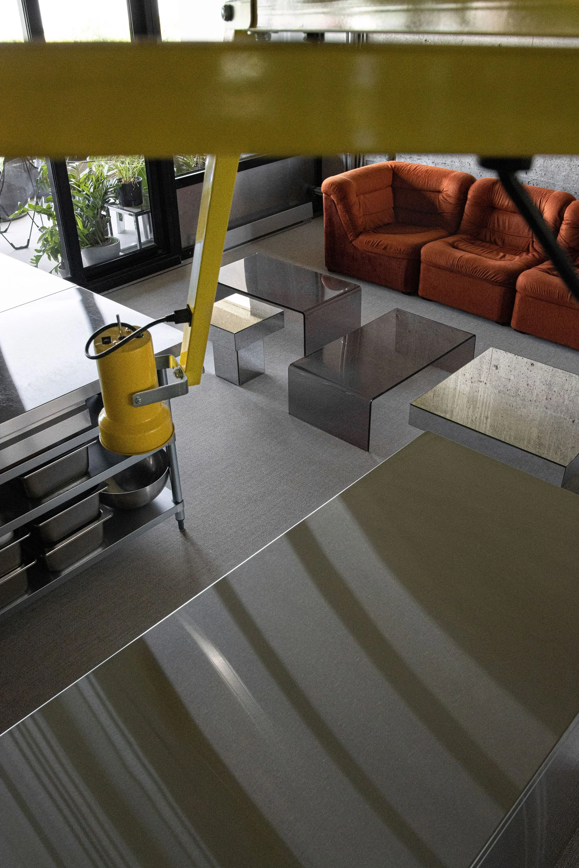 An industrial living room features a modular burnt orange sofa, several translucent smoky glass coffee tables, and a yellow industrial lamp.