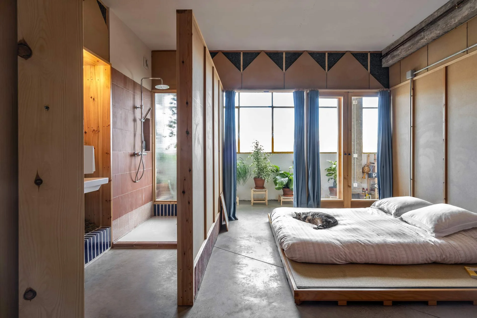 A minimalist loft bedroom features a low wooden platform bed, a concrete floor, and a pine partition wall separating the sleeping space from an open shower.