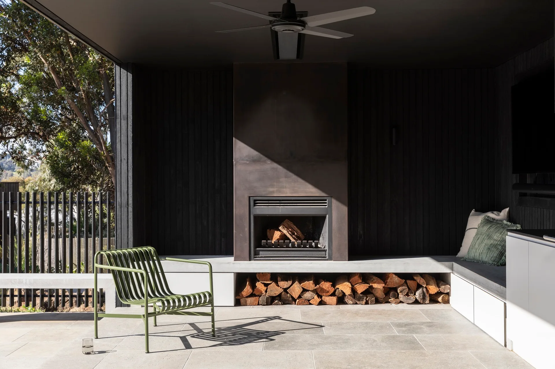 A modern covered patio features a black vertical wood slat wall, an inset fireplace, a low concrete bench with wood storage, and a sage green metal chair.
