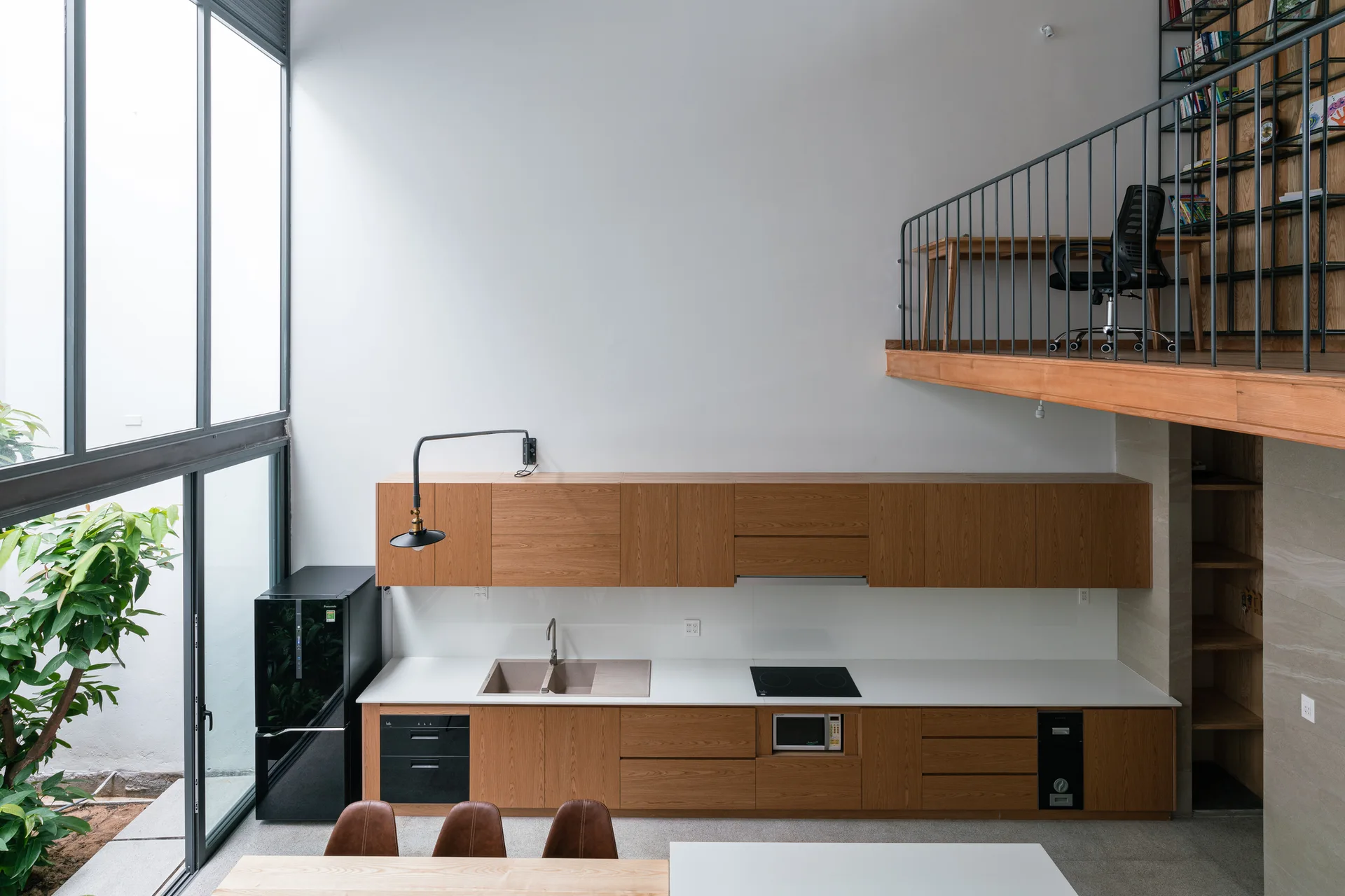 A modern kitchen with wood cabinets and white countertops sits below a second-floor loft workspace with black metal railings.
