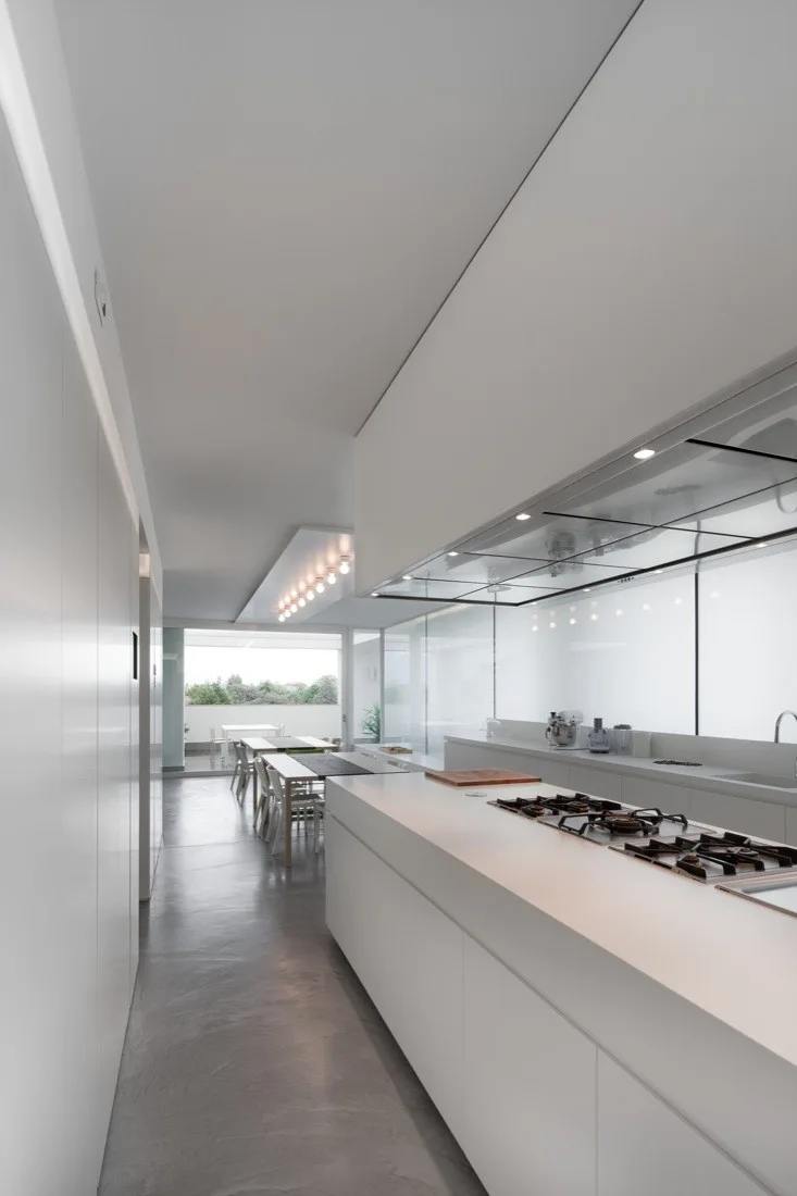 A long minimalist white kitchen features a large central island with a cooktop, seamless floor-to-ceiling cabinets, and a dining table in the background.