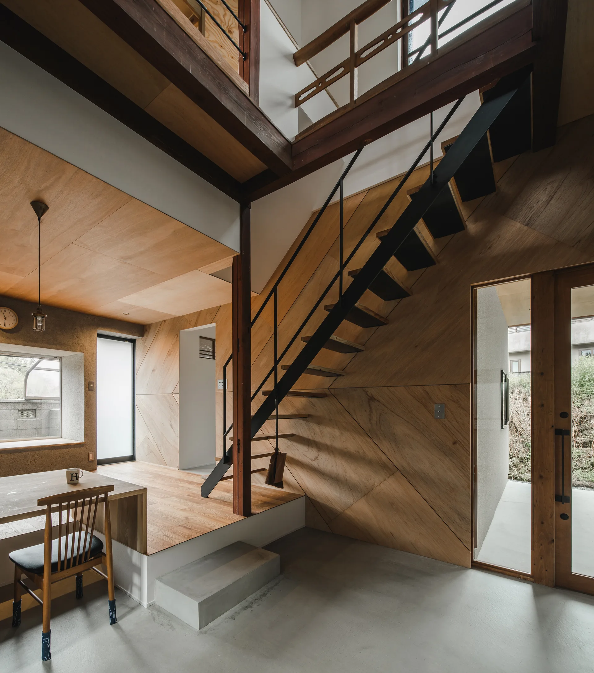 An entryway features a concrete floor, a raised wood platform, and a black metal staircase against a geometric wood-paneled wall.