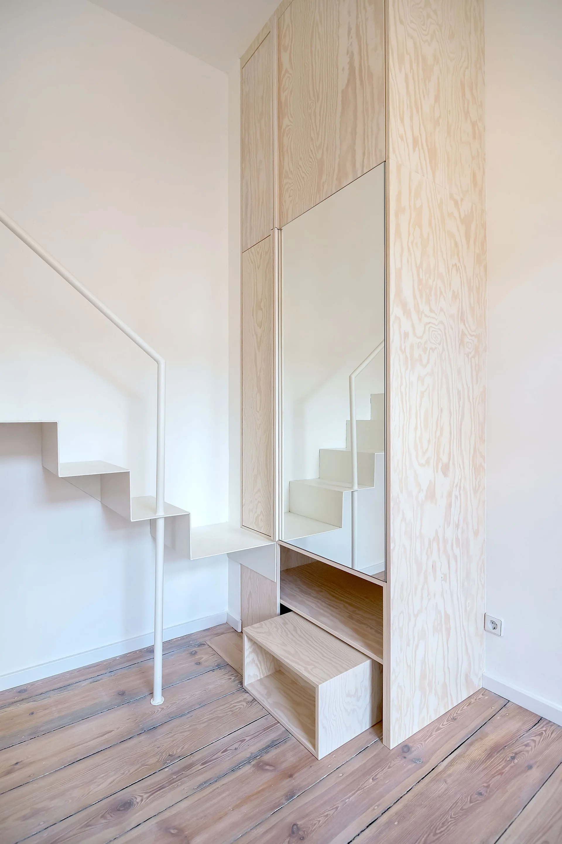 A minimalist white metal staircase stands next to a tall custom plywood cabinet with an integrated mirror on light wood floors.