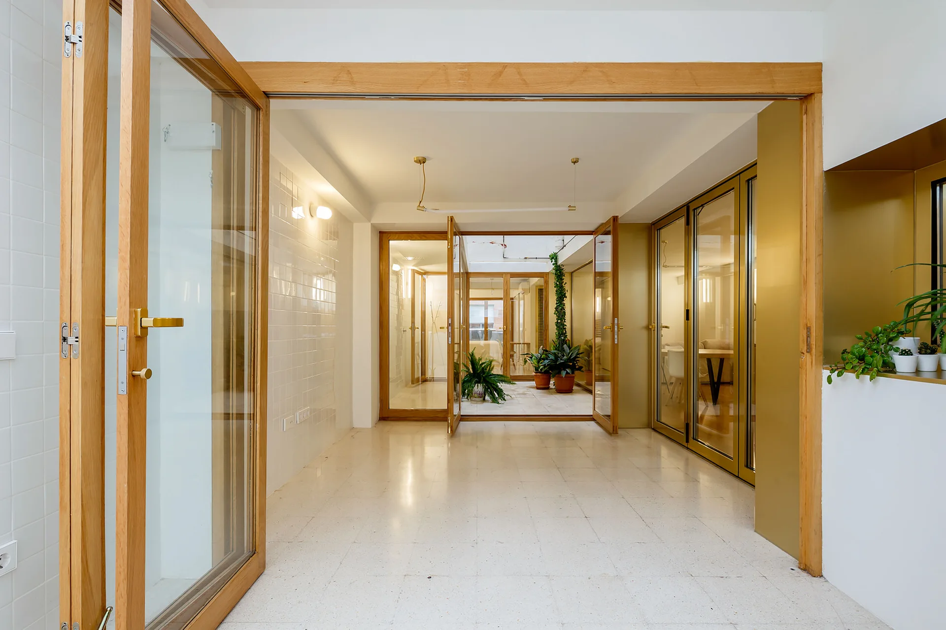 A bright transitional hallway featuring glossy white tiled walls, wood-framed glass doors, and a view into a small courtyard with green plants.