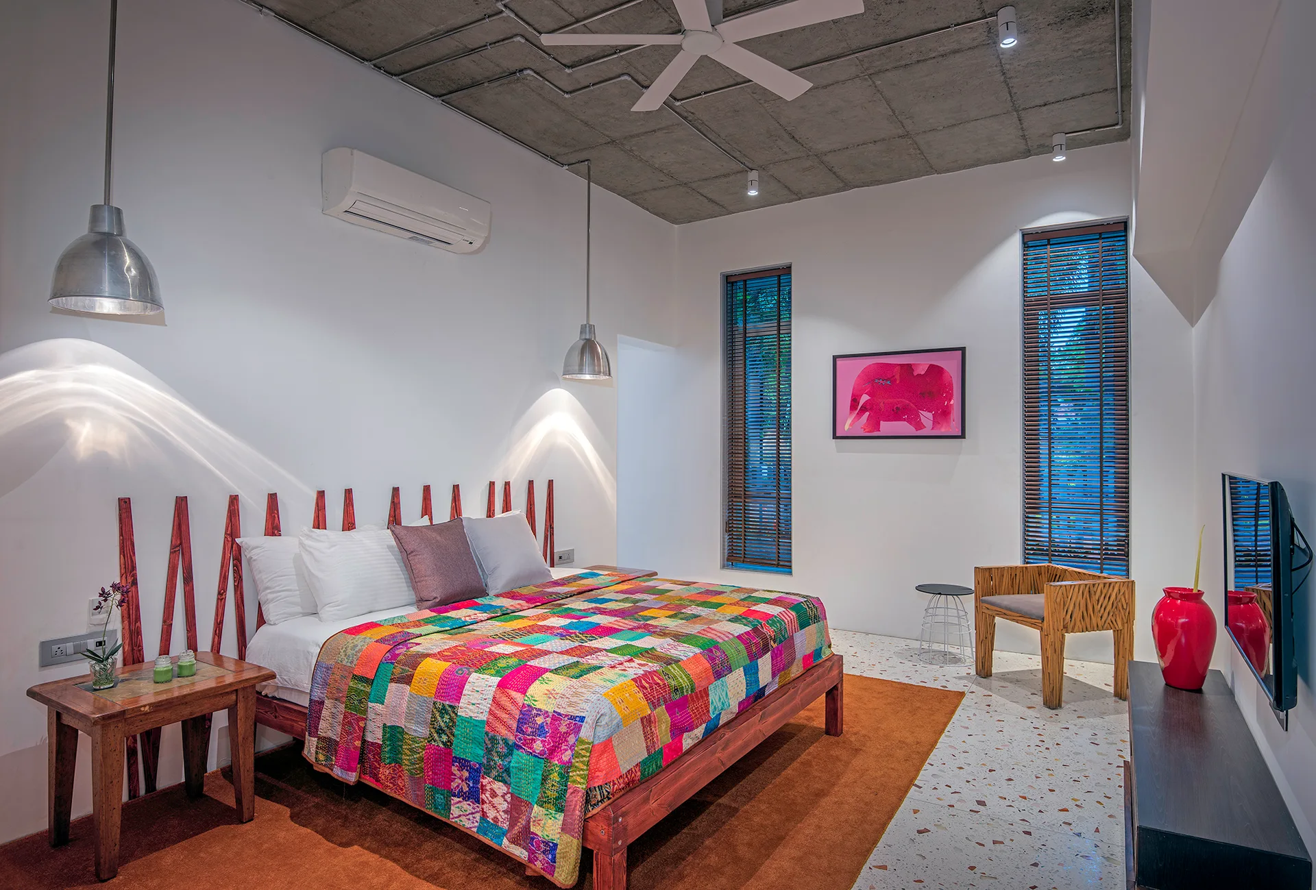 A modern industrial bedroom featuring an exposed concrete ceiling, a colorful patchwork quilt on a wood bed, and vertical wooden slats as a headboard.