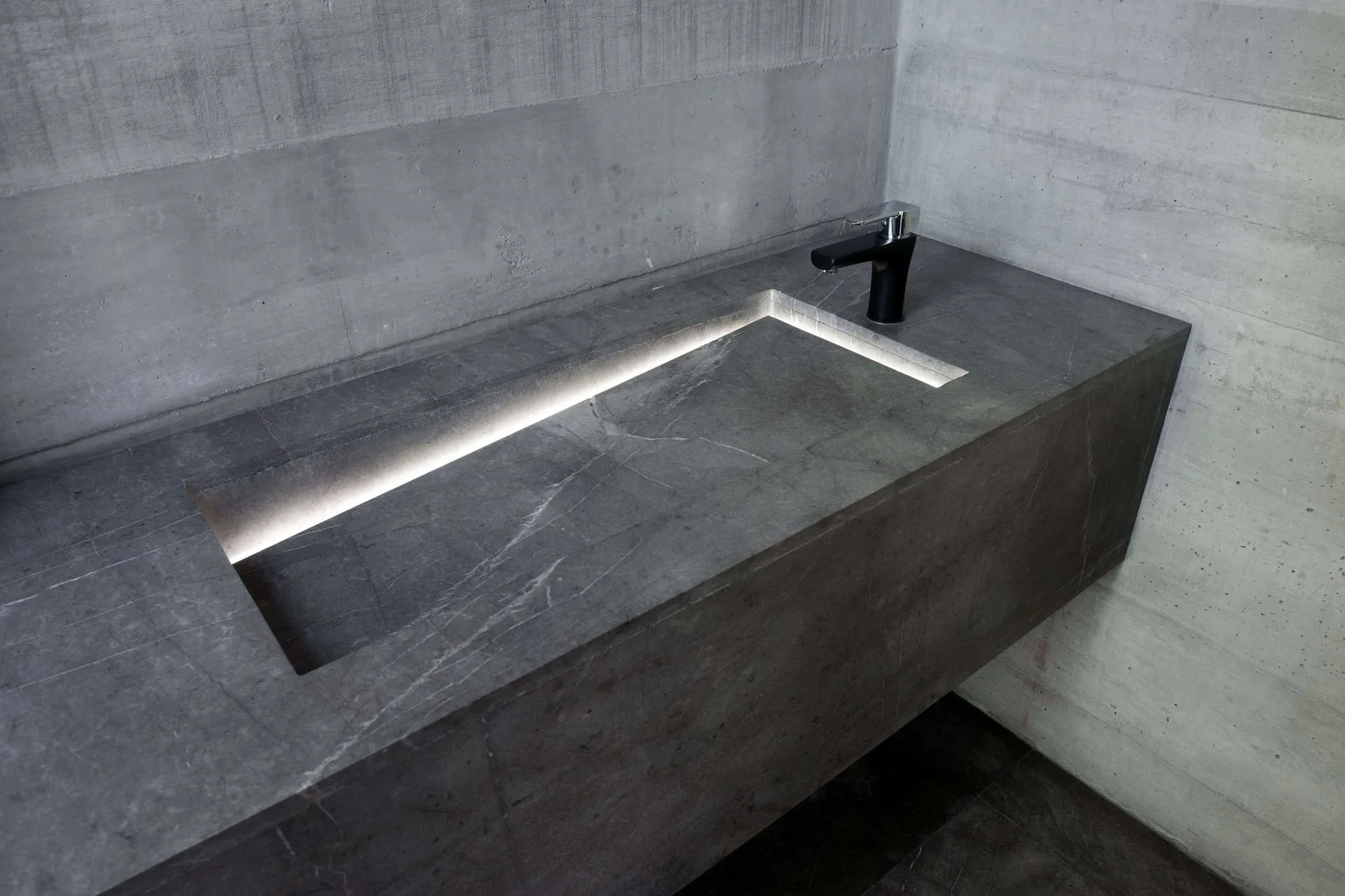 A modern wall-mounted gray stone vanity features an integrated sink illuminated by a recessed LED light against raw concrete walls.