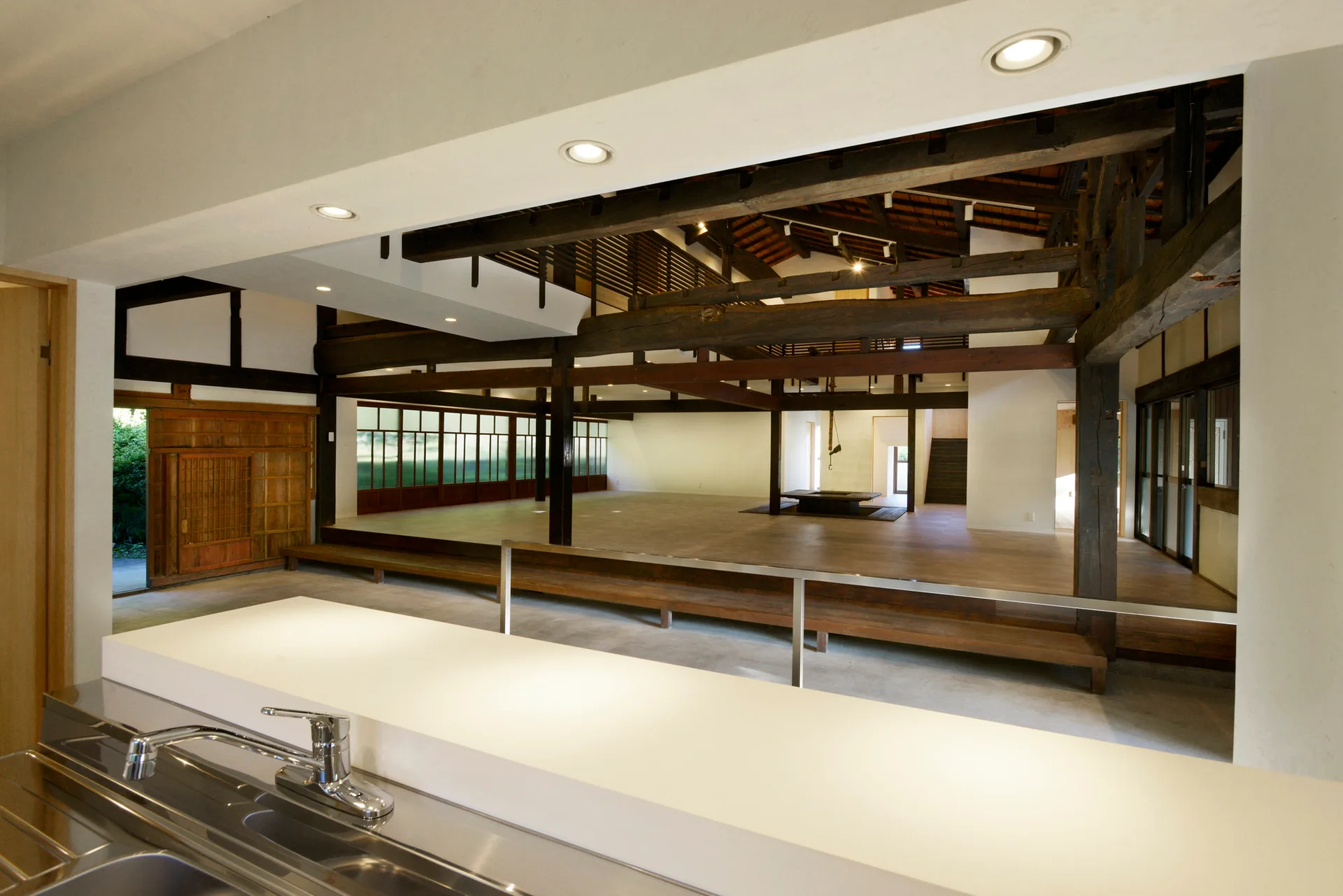 A modern white kitchen counter overlooks a large, open living area featuring a raised wooden platform and exposed dark timber ceiling beams.