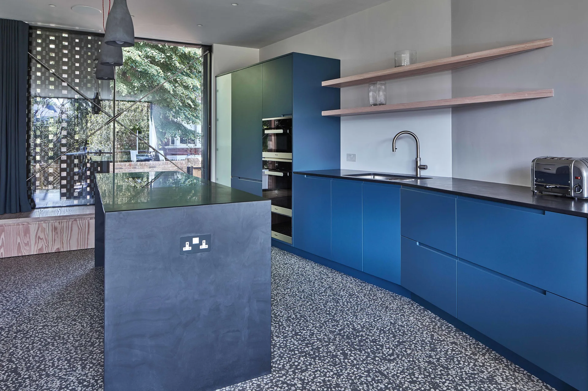 A modern kitchen featuring matte blue flat-front cabinets, a dark grey island, terrazzo flooring, and light wood floating shelves.