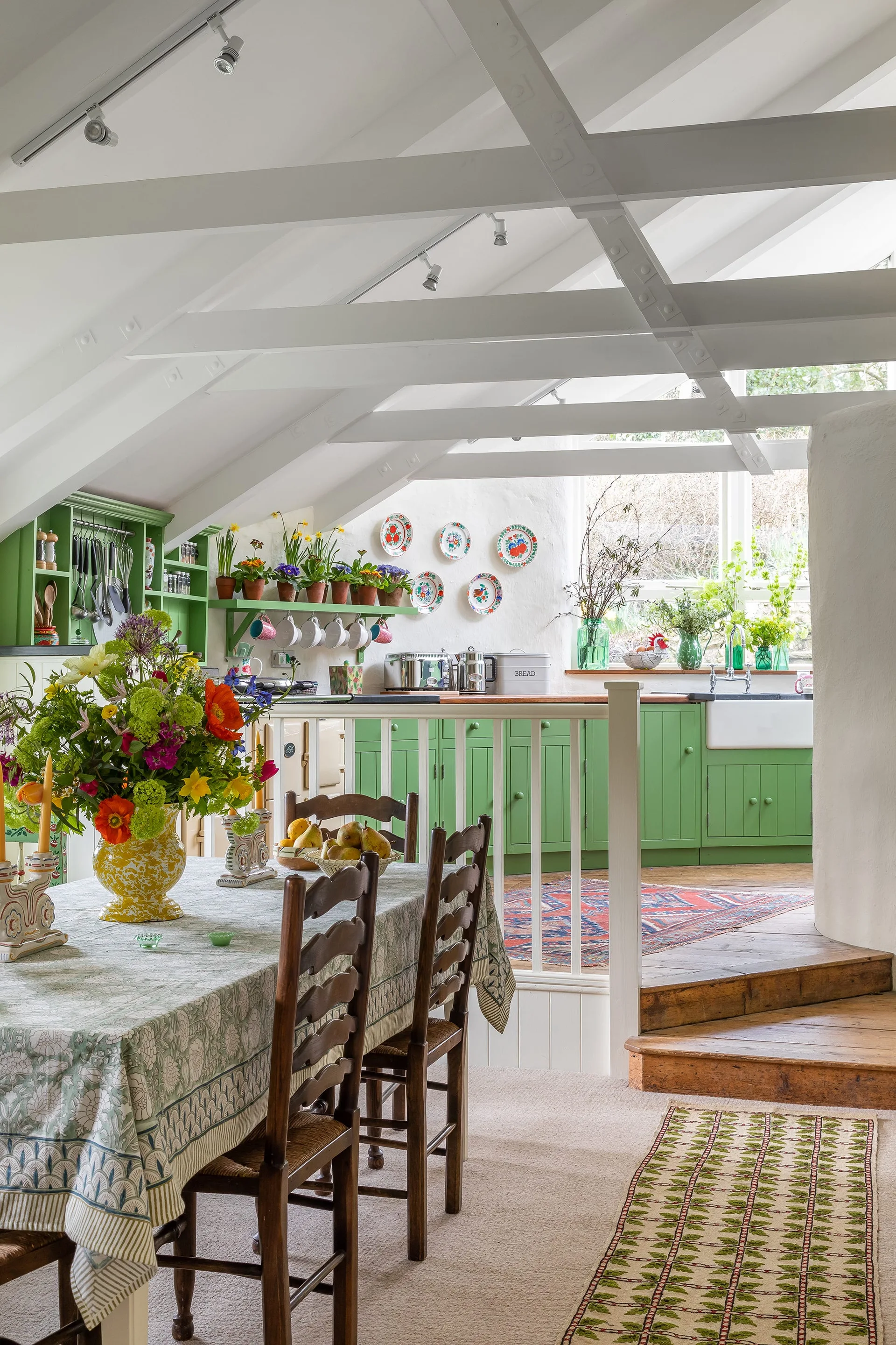 A split-level cottage interior features a dining table with wooden chairs in the foreground and an elevated green kitchen with white ceiling beams.