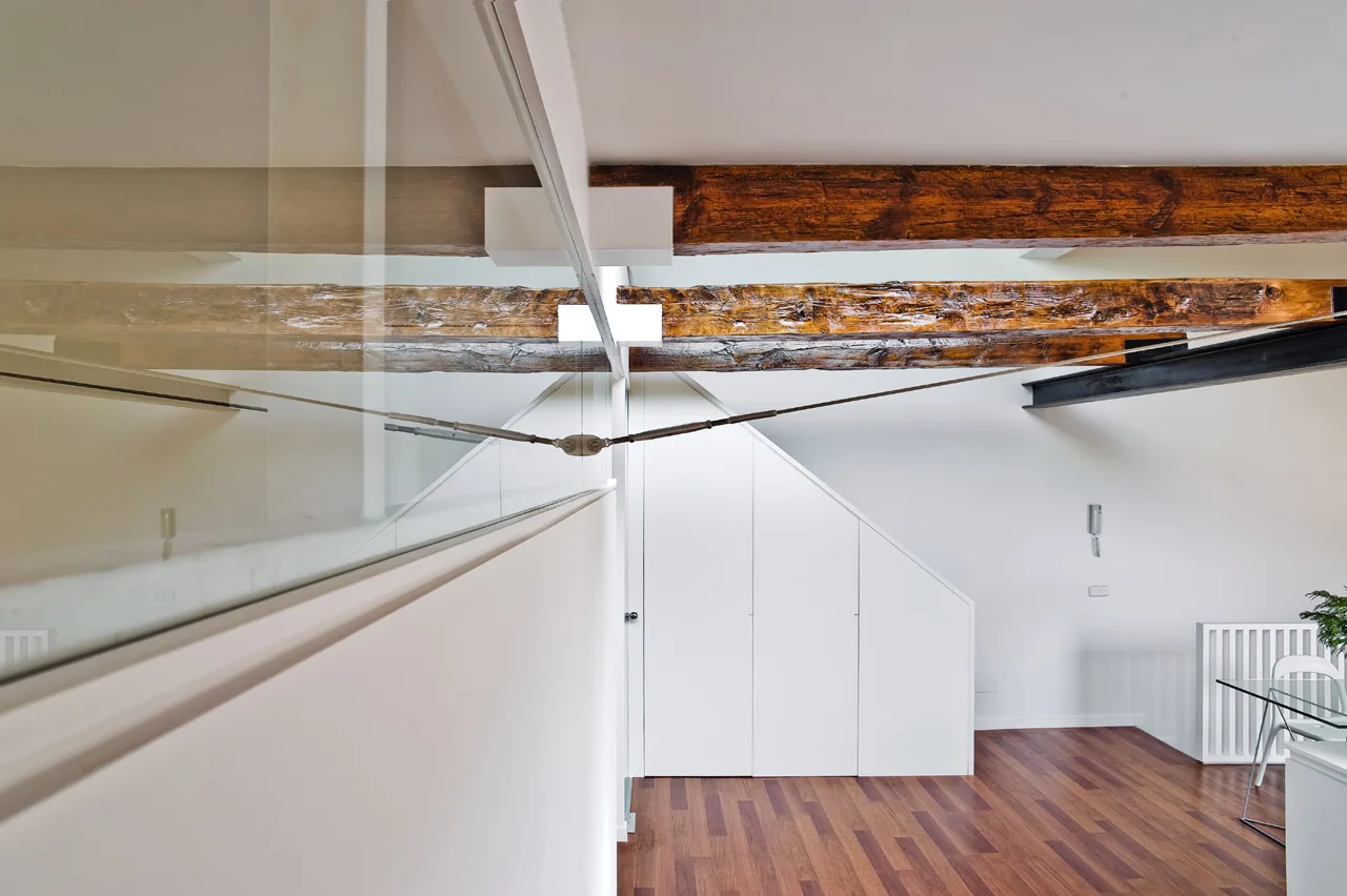 A modern interior space with exposed rustic wooden ceiling beams, a glass partition wall, and white built-in storage under a sloped ceiling.