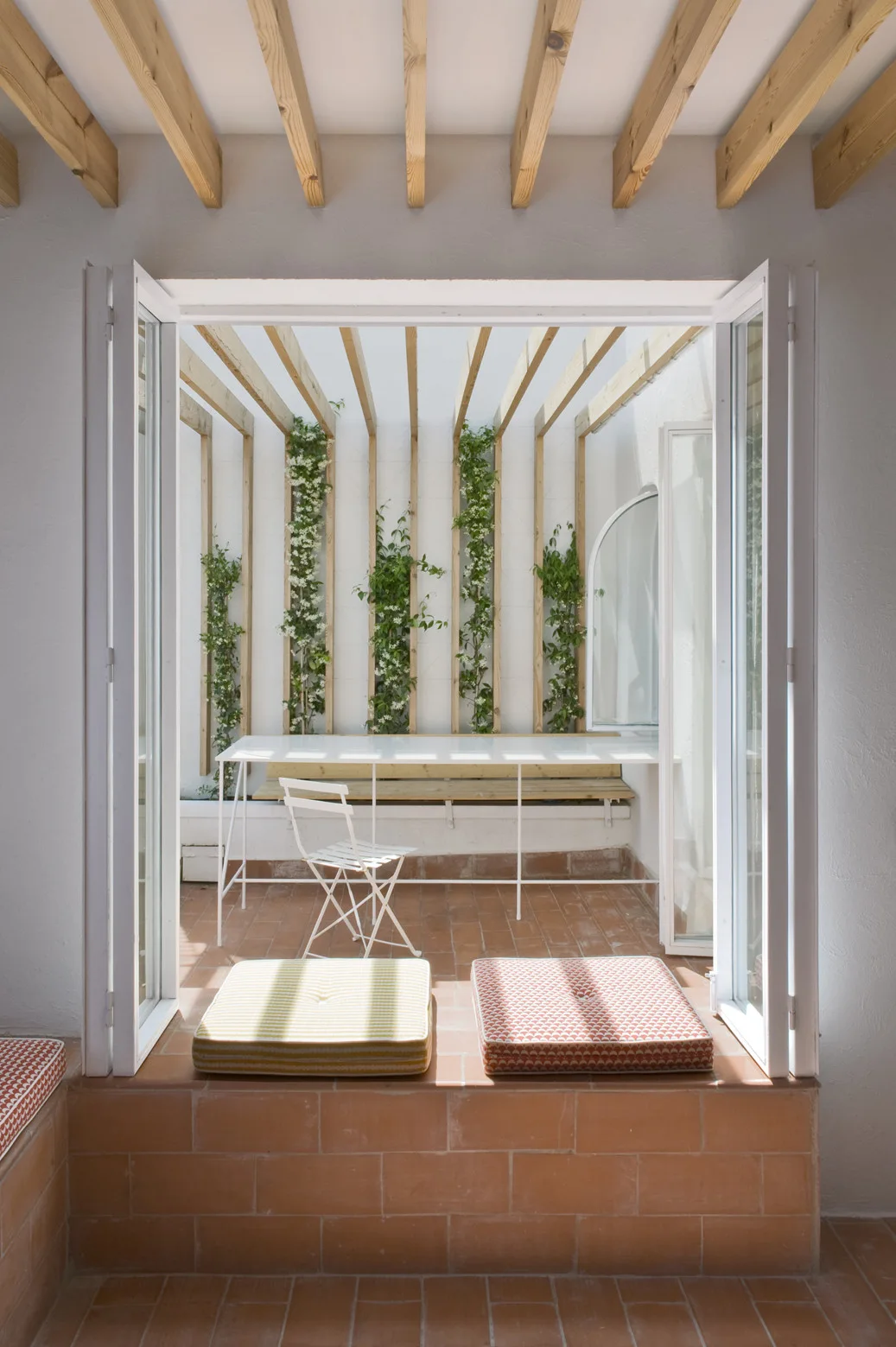 An outdoor patio with terracotta tiles features a white table, a folding chair, and vertical wood trellises with climbing green plants.