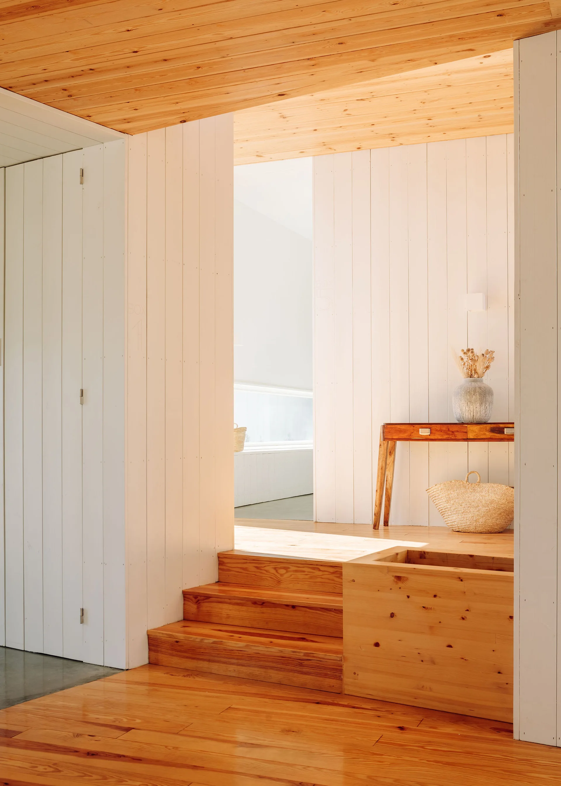 A light-filled hallway features vertical white wood paneled walls, a natural wood staircase, a matching ceiling, and a simple wooden console table.