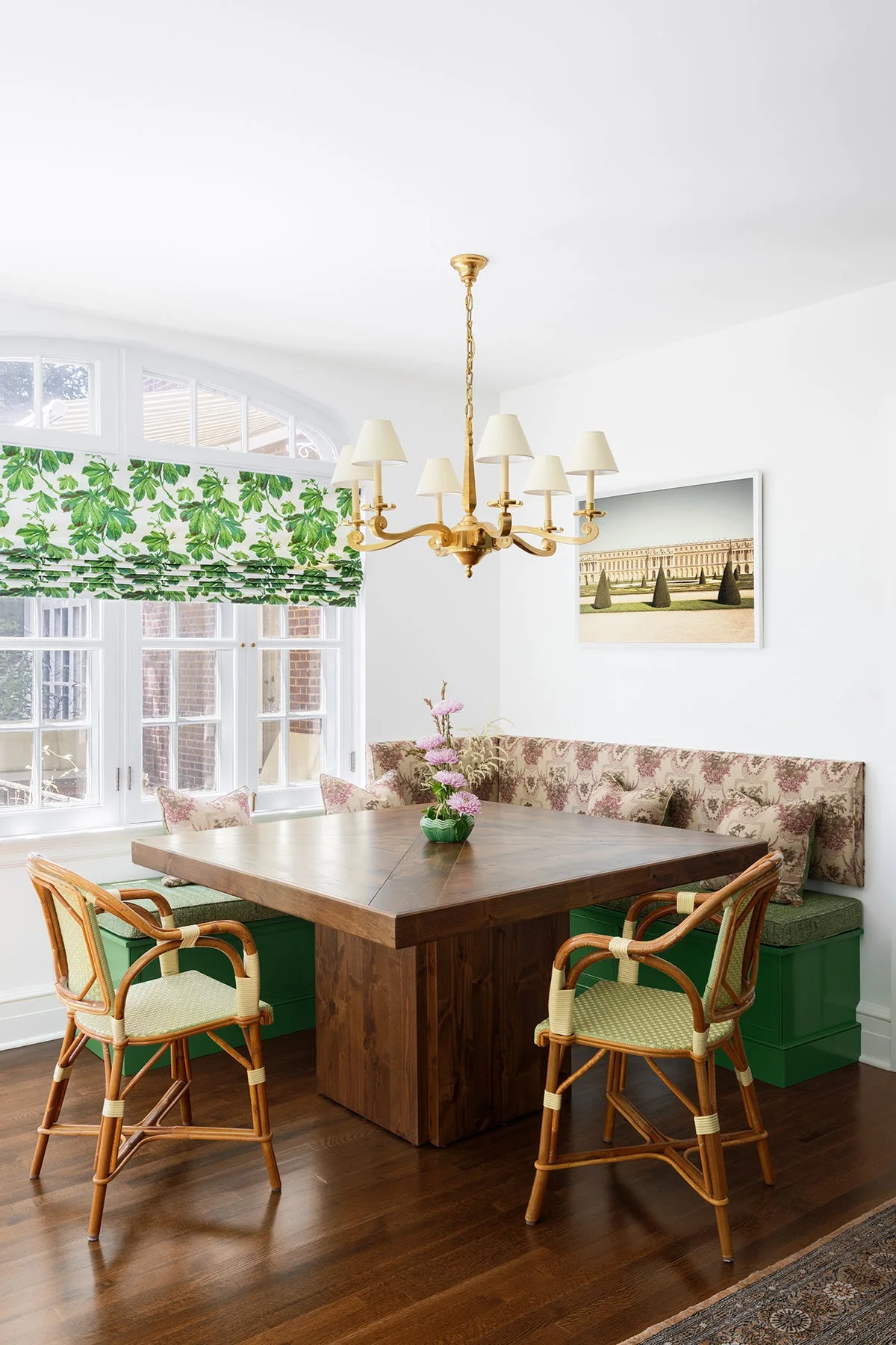 A bright dining nook features a square wooden table surrounded by a green built-in banquette, two rattan chairs, and a brass chandelier.