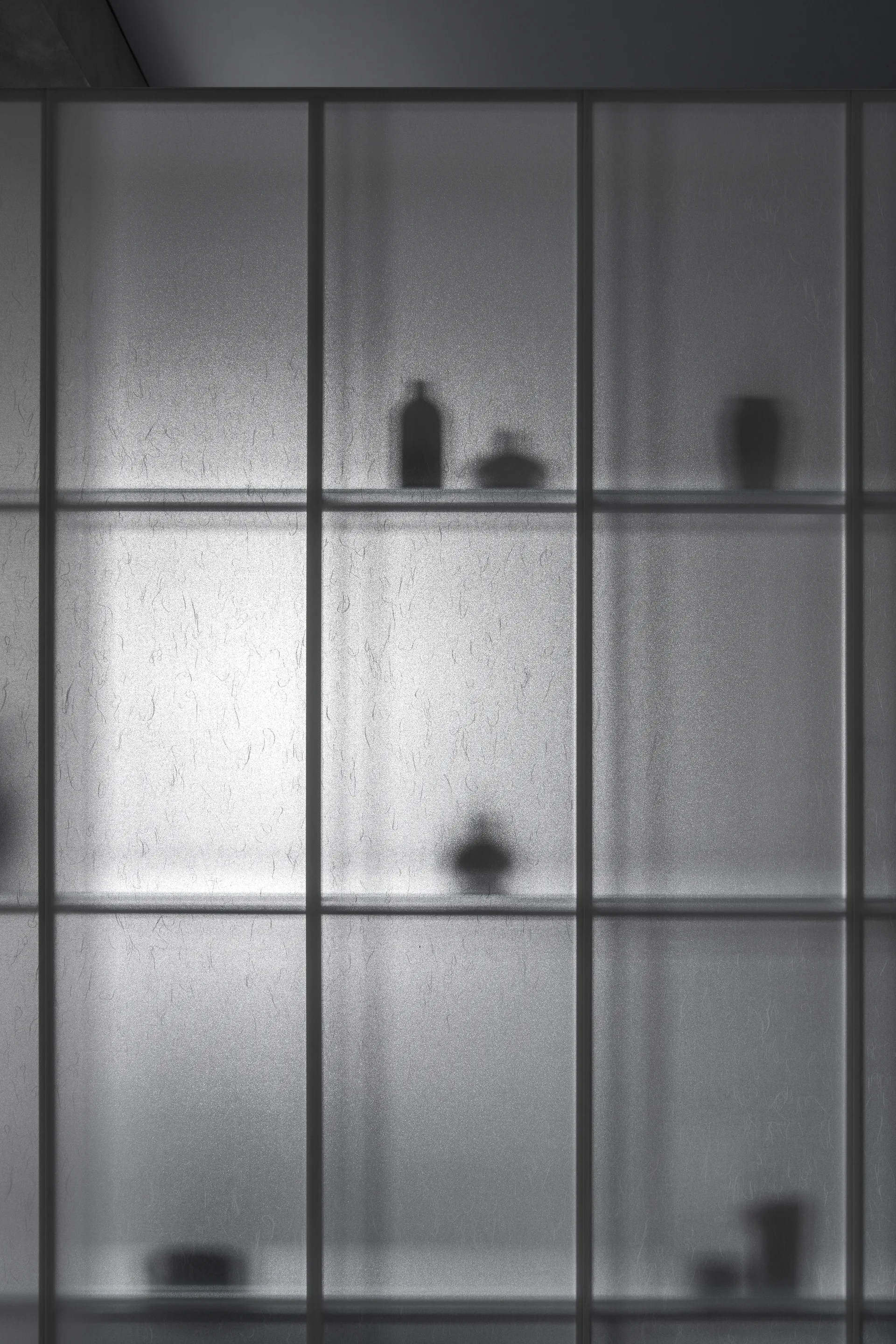 A close-up of a minimalist storage cabinet featuring frosted glass grid doors that reveal the soft silhouettes of vases and bottles on internal shelves.