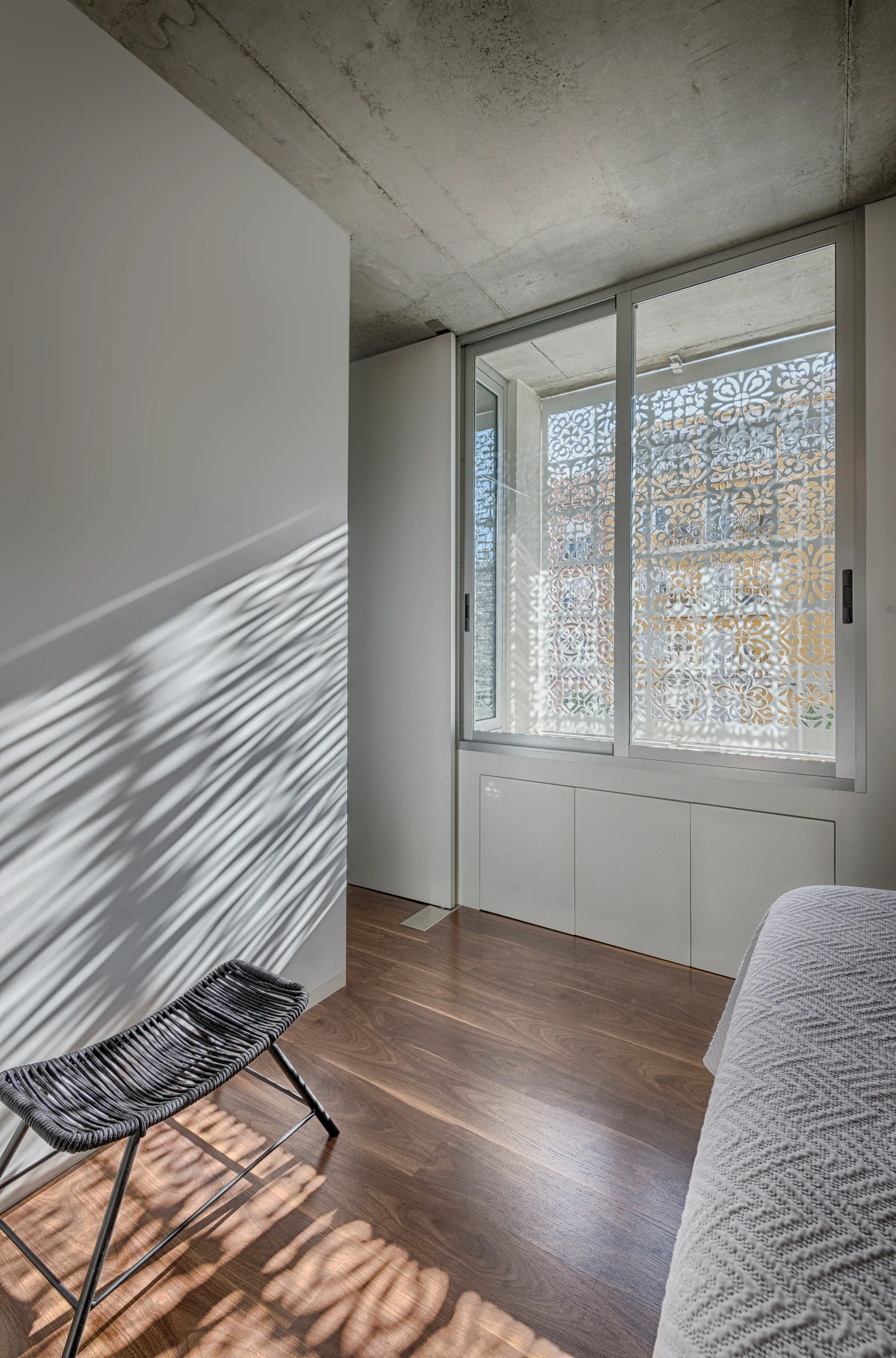 A minimalist bedroom features dark wood flooring, a raw concrete ceiling, a black woven stool, and a window with an ornate geometric pattern screen.