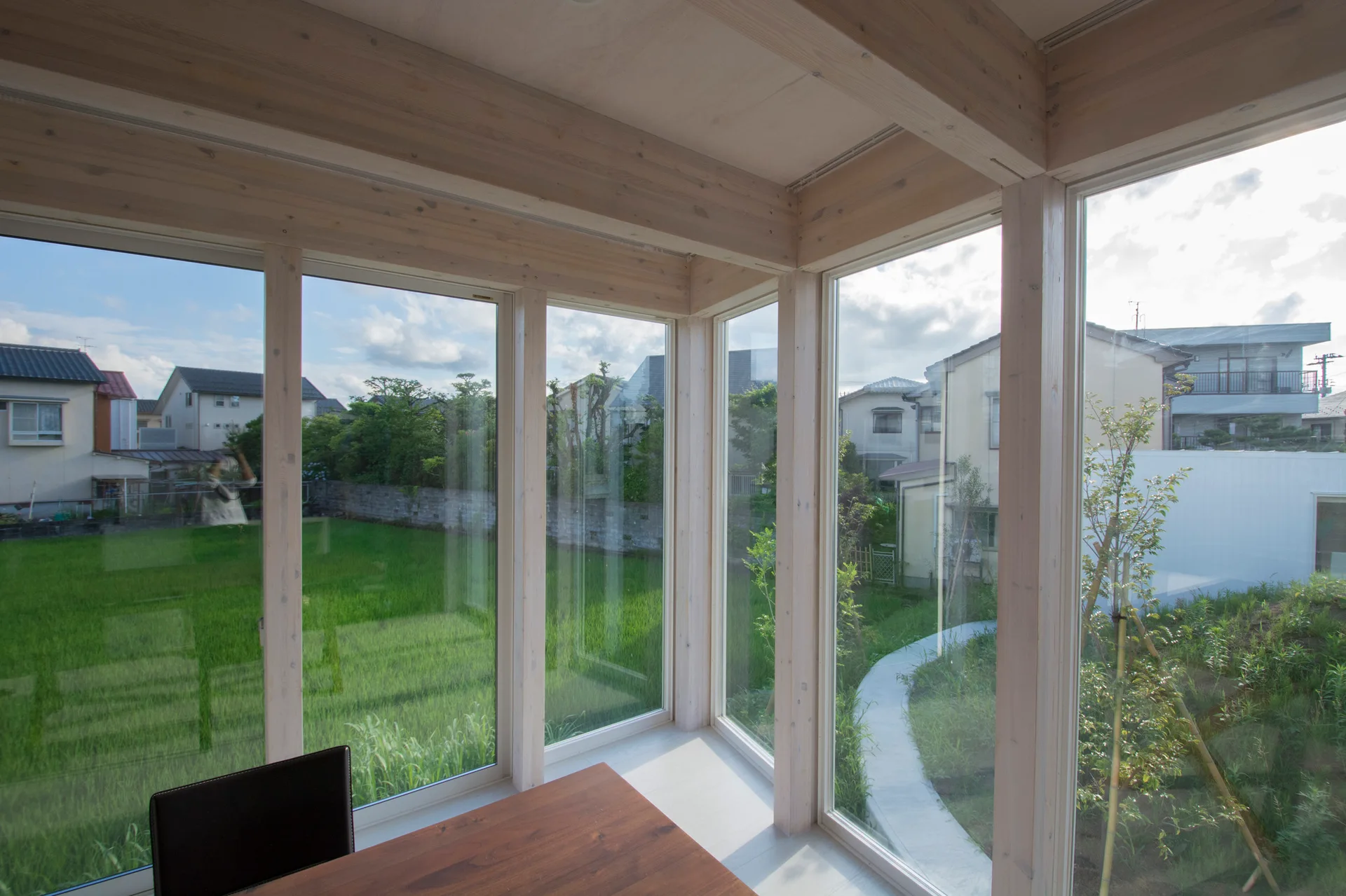 A minimalist interior with light wood ceiling beams, structural timber framing, and floor-to-ceiling windows showing a residential yard.