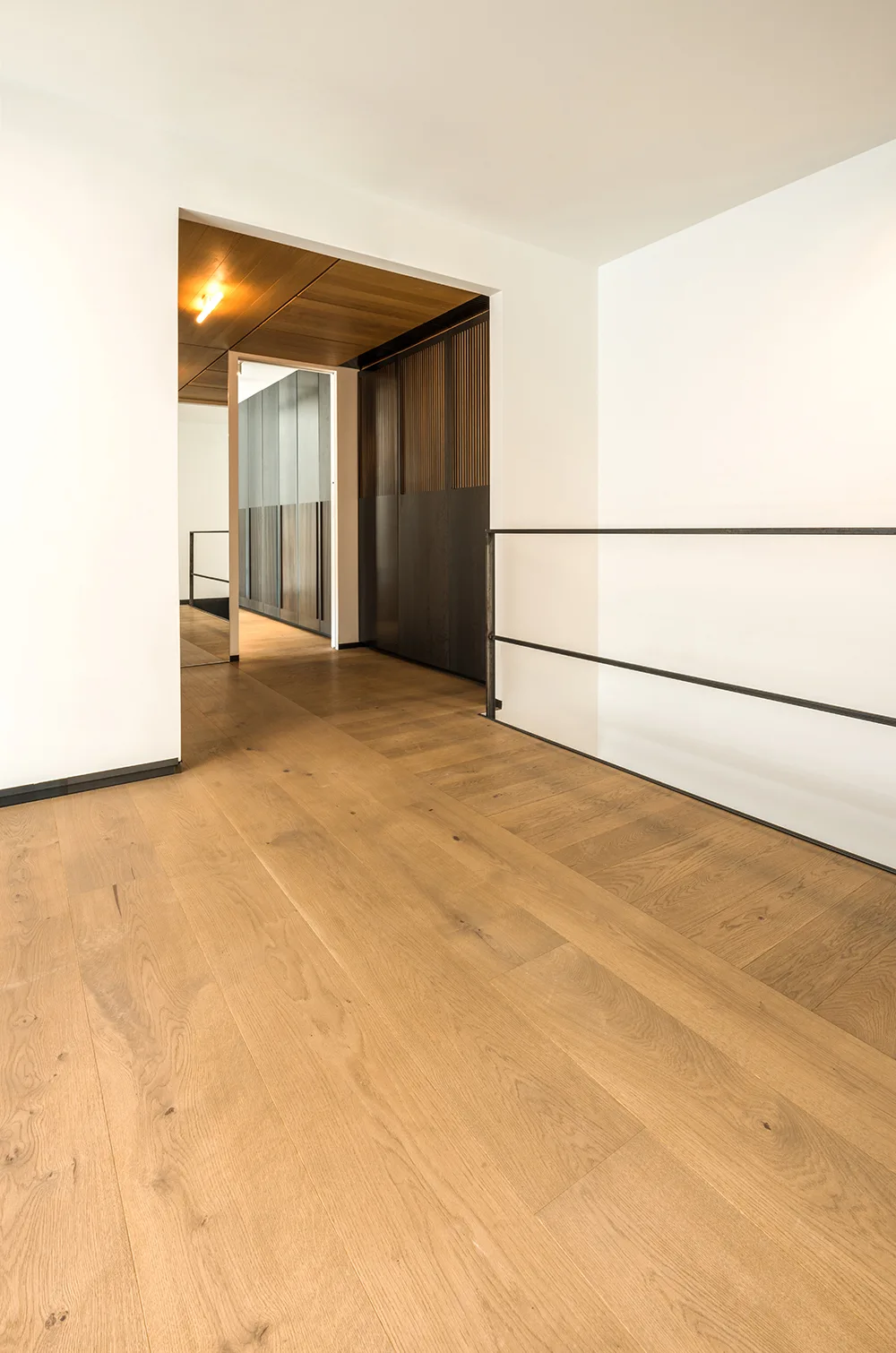 A minimalist hallway features light oak flooring, bright white walls, a thin black metal railing, and a dark wooden slatted accent wall.