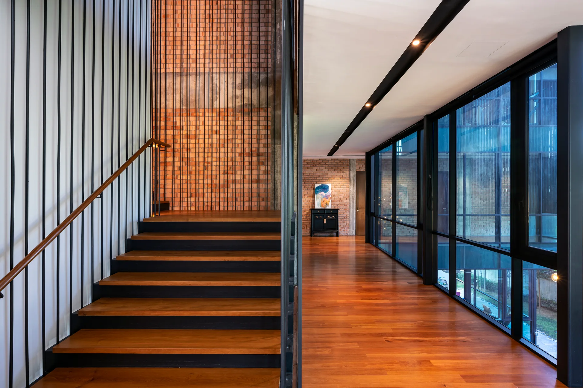 A modern wooden staircase with vertical black metal railings stands next to a wide hallway featuring polished wood floors, exposed brick, and large windows.