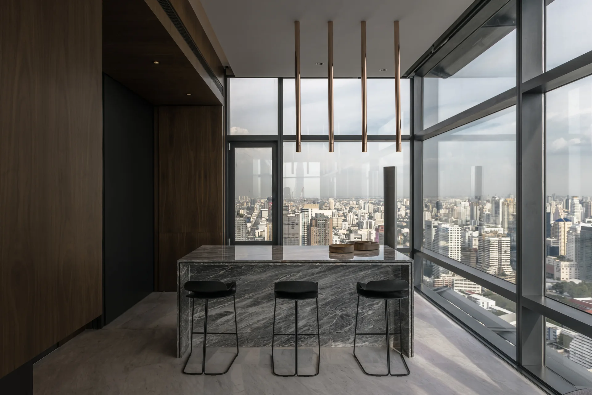 A grey marble waterfall island with three black bar stools sits in front of floor-to-ceiling windows overlooking a panoramic city skyline.