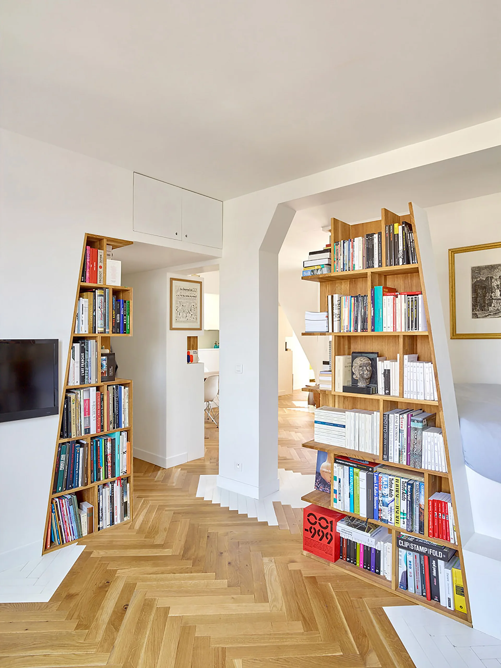 Custom-built angled wooden bookshelves frame a white archway in a modern room with oak herringbone parquet floors.