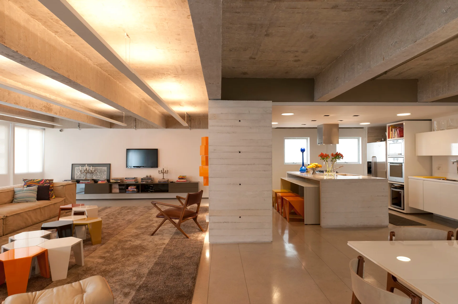 An open-plan living room and kitchen separated by a central concrete pillar, featuring a beige sofa, wooden armchair, and modern cabinetry.