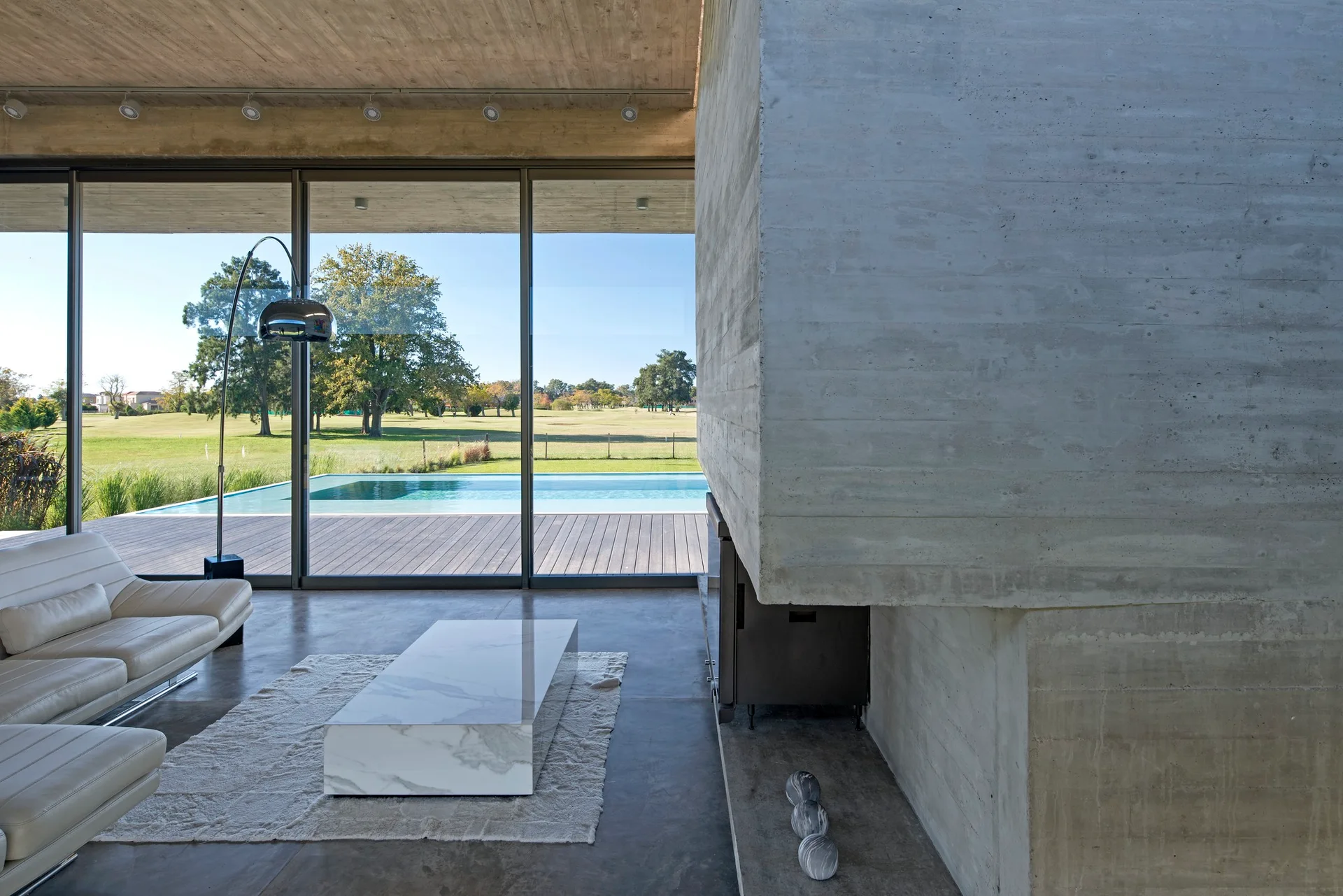 A minimalist living room with a raw concrete fireplace, a low white marble coffee table, and large glass doors opening to an outdoor pool.